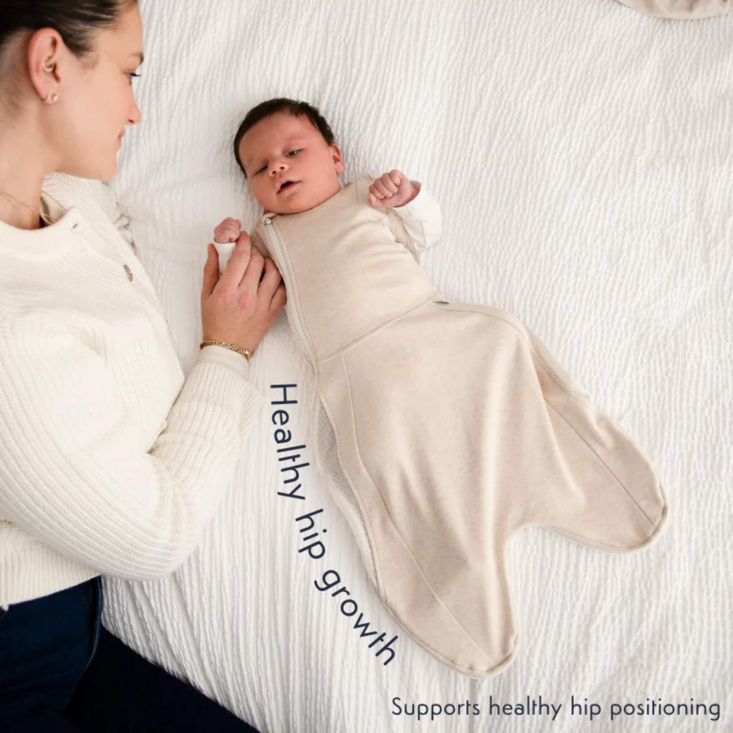A baby lying beside an adult while wearing the swaddle bag, illustrating the hip-friendly shape that supports natural leg movement.