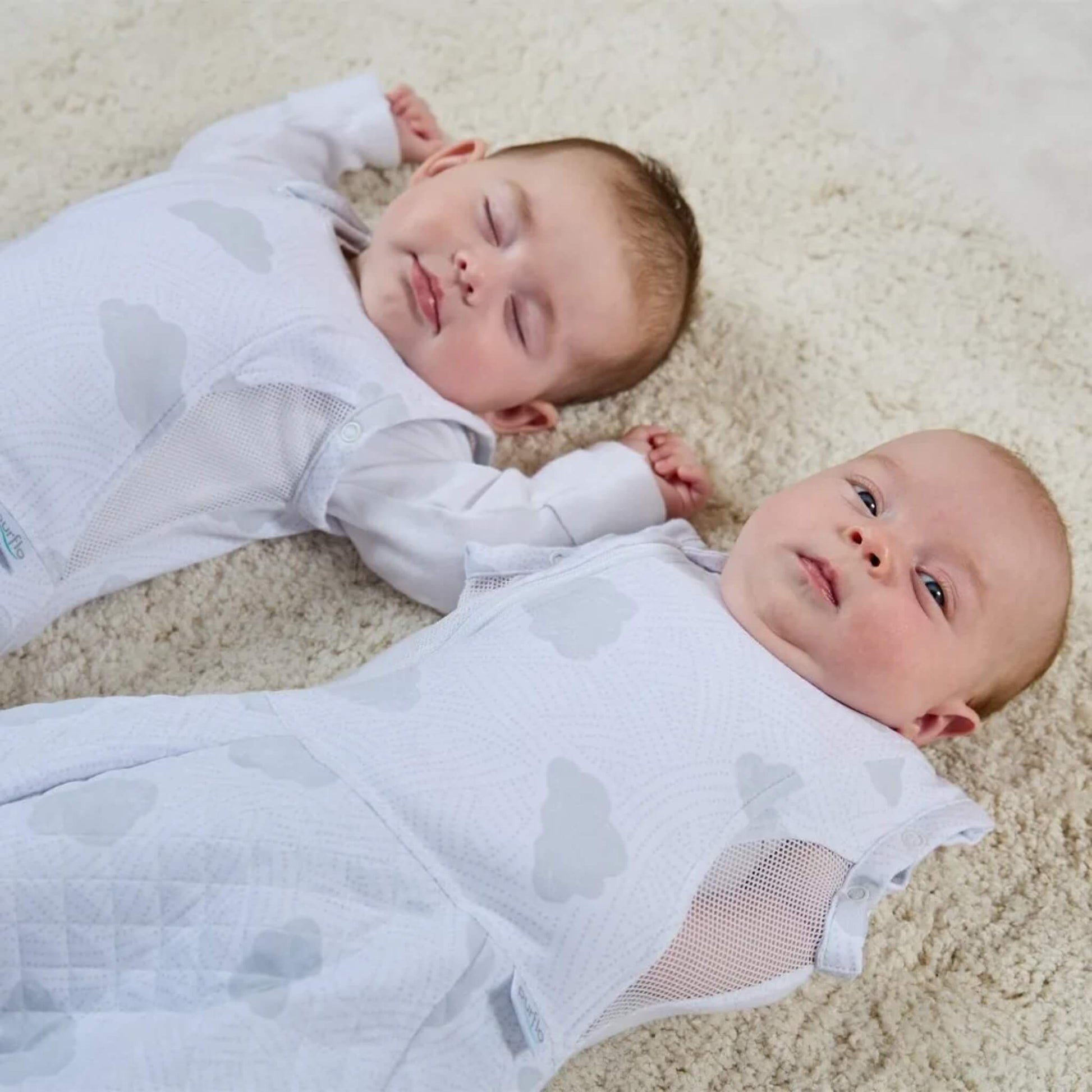 A close view of two babies lying on a soft rug in matching cloud-print sleep bags, showing breathable mesh side panels and one in relaxed arm positioning and other baby arms in.
