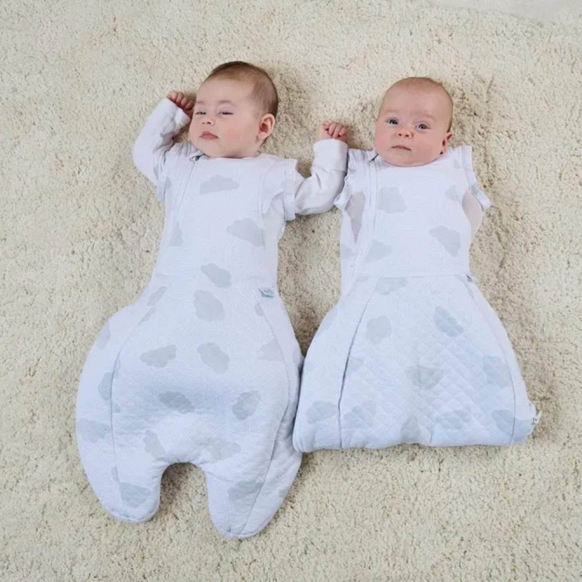 Two babies lying side by side on a soft rug, both wearing cloud-print sleep bags, one with arms fully enclosed inside the bag and the other with arms released through the armholes.