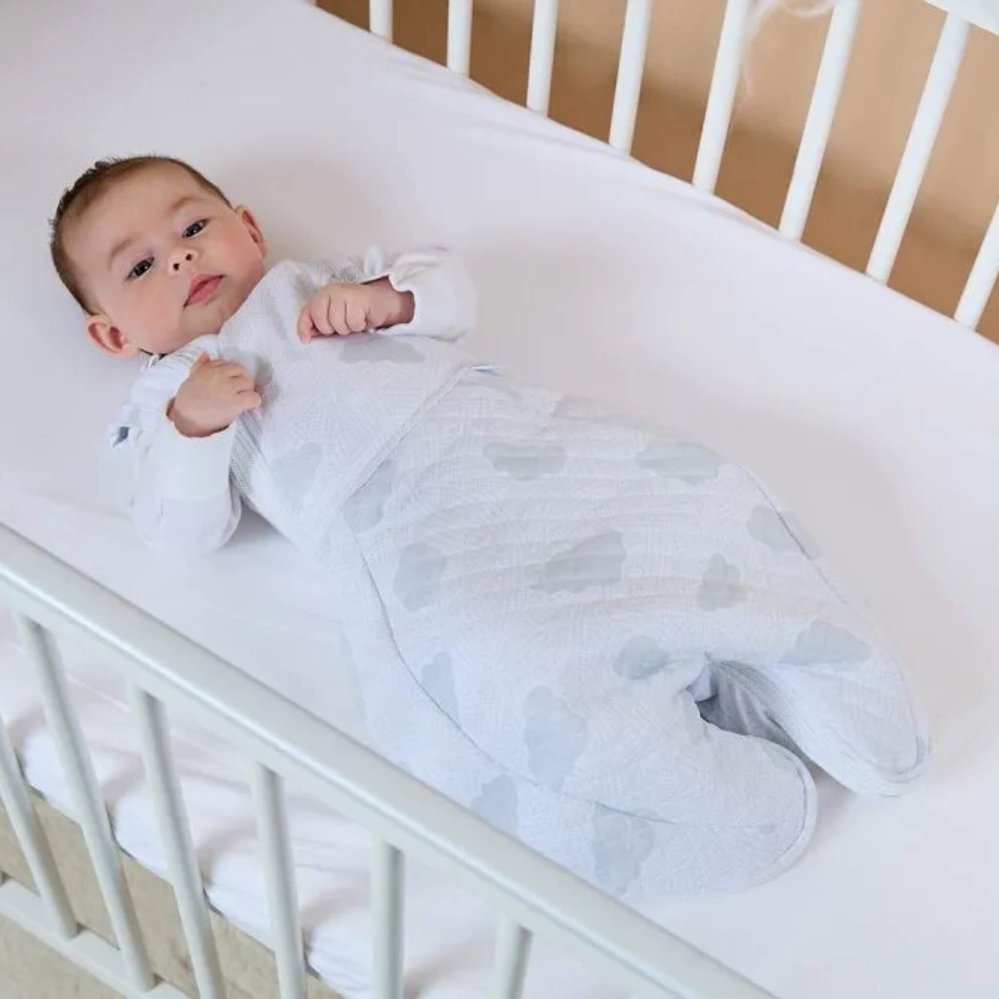 Baby lying in cot wearing swaddle sleep bag with arms out, cloud print visible across chest and legs.