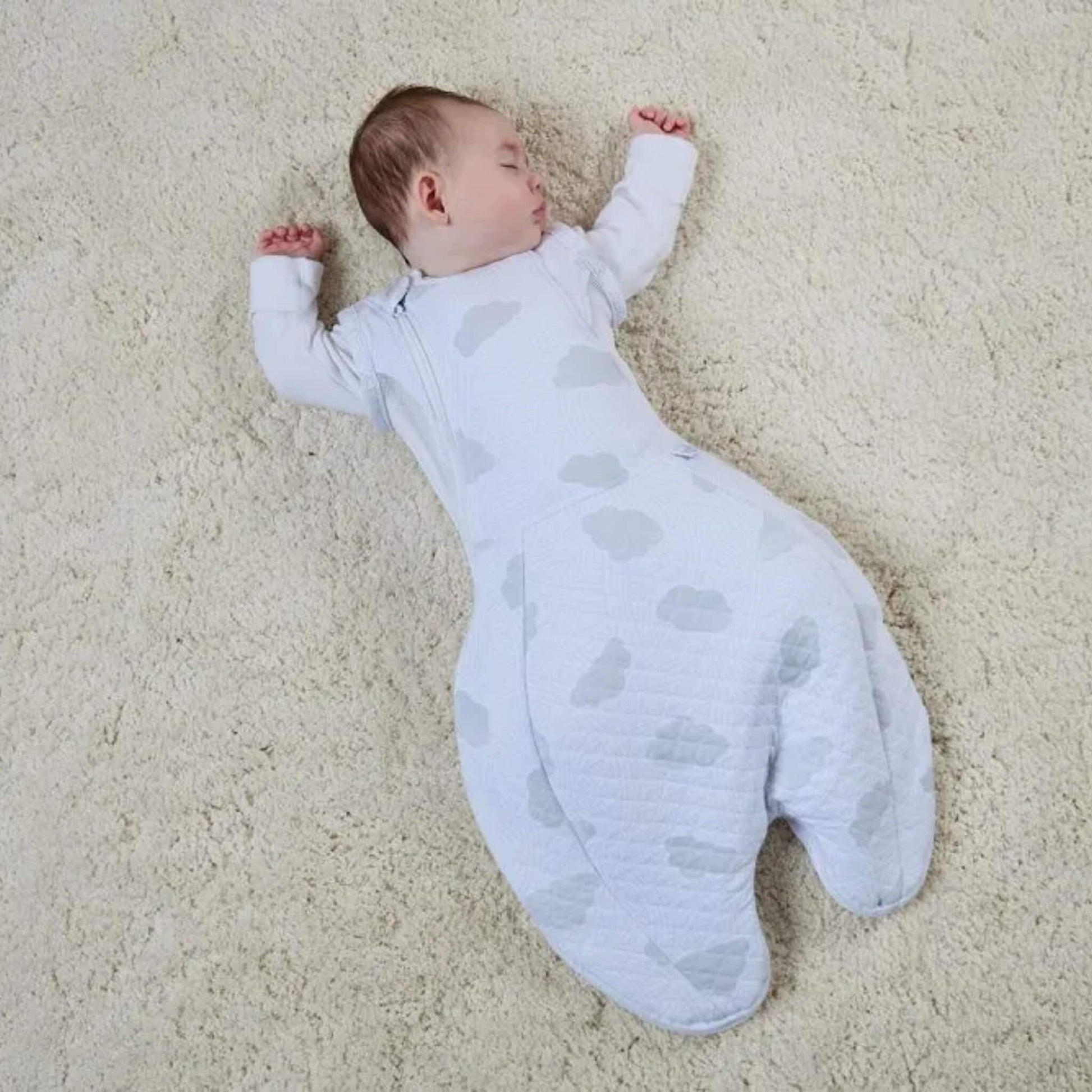 Baby sleeping on soft surface wearing swaddle sleep bag with relaxed arms-out position and extended legs.