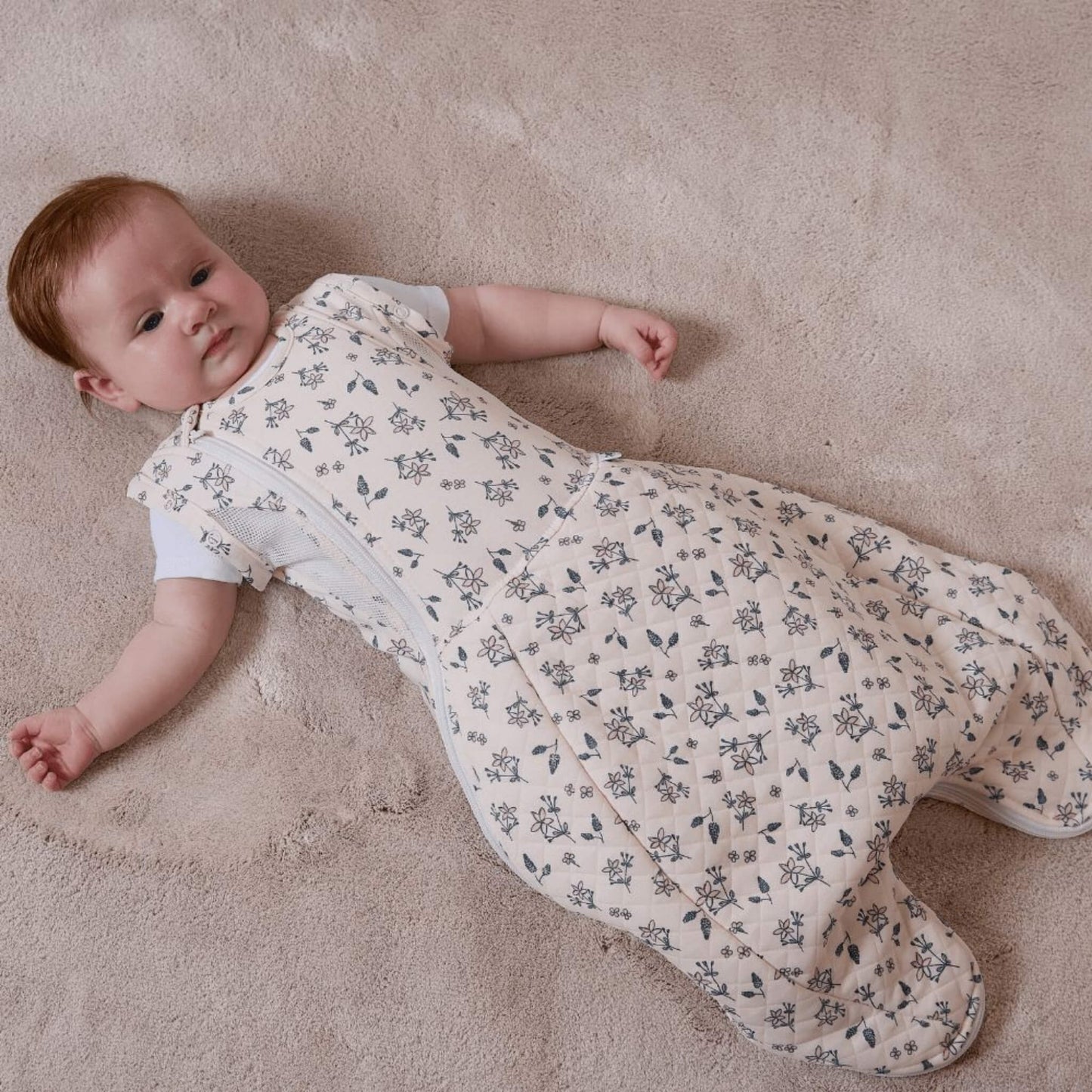 A baby lying on a soft carpet wearing a swaddle to sleep bag with arms out, showing the transition from swaddling to a standard sleep bag.