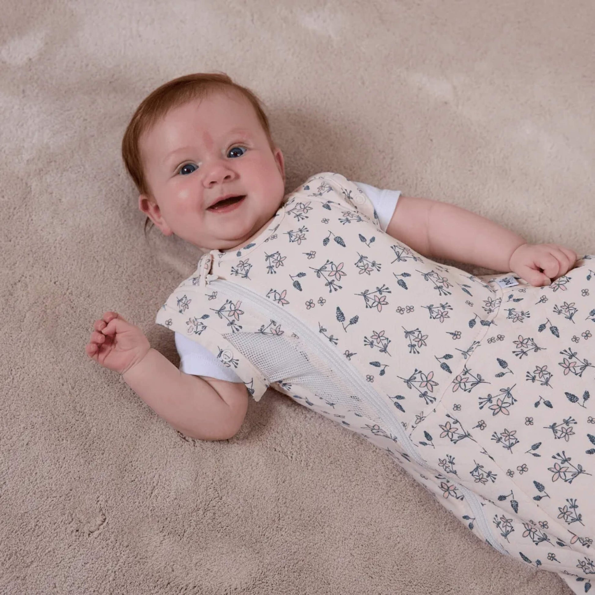 A baby lying on a soft carpet wearing a swaddle to sleep bag with arms out, highlighting the breathable mesh side panel and front zip.