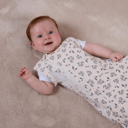 A baby lying on a soft carpet wearing a swaddle to sleep bag with arms out, highlighting the breathable mesh side panel and front zip.