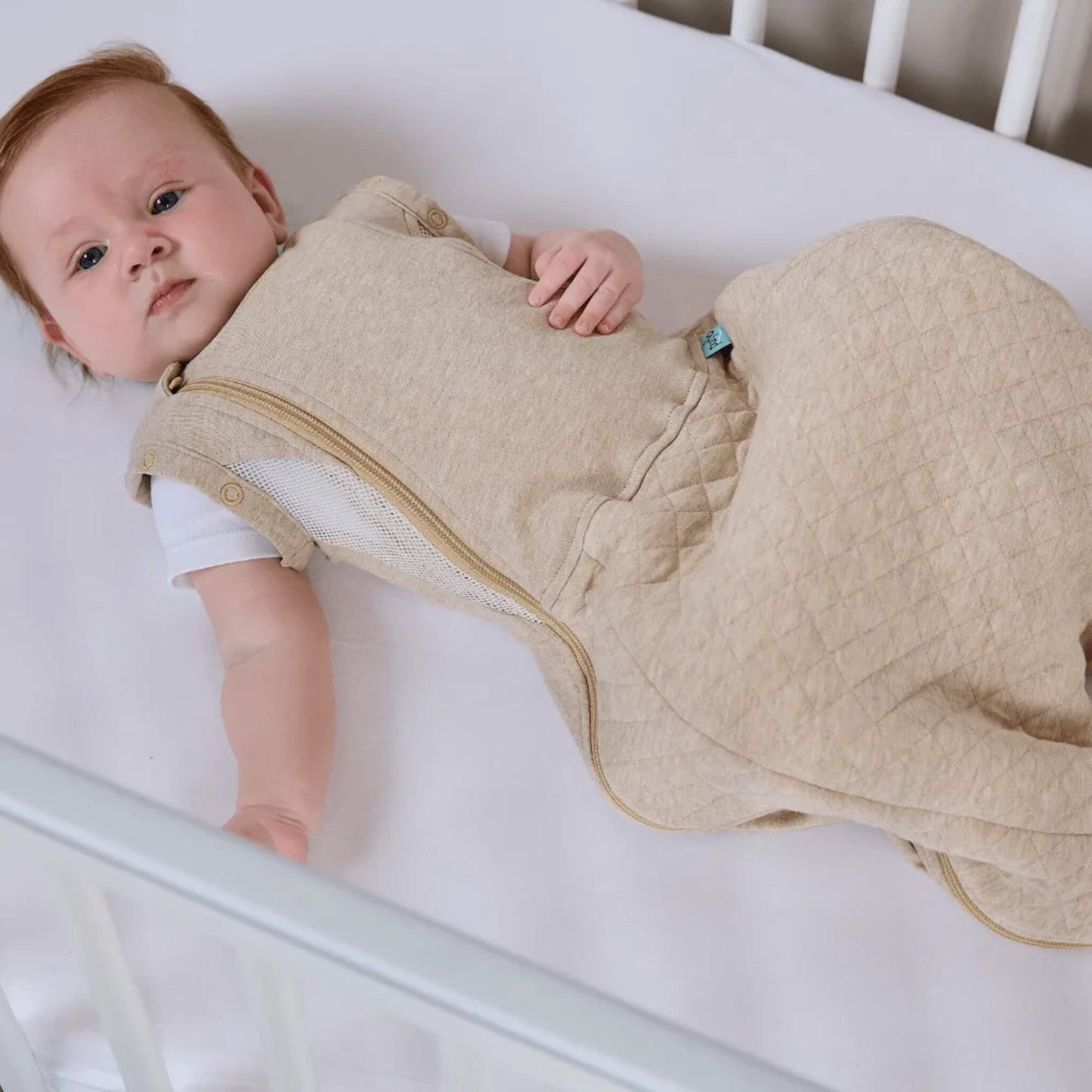 A baby resting in a cot wearing a beige sleep bag with a quilted lower section visible.
