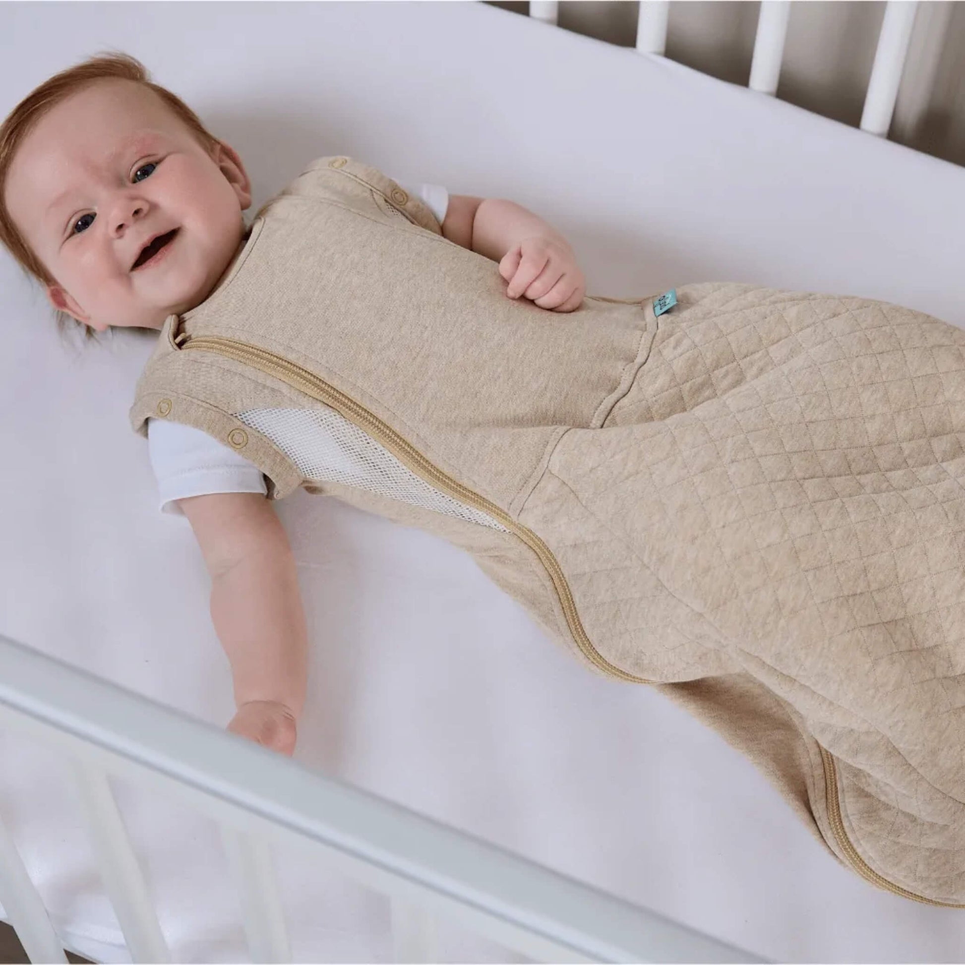 A baby lying on a mattress wearing a beige sleep bag with arms free.