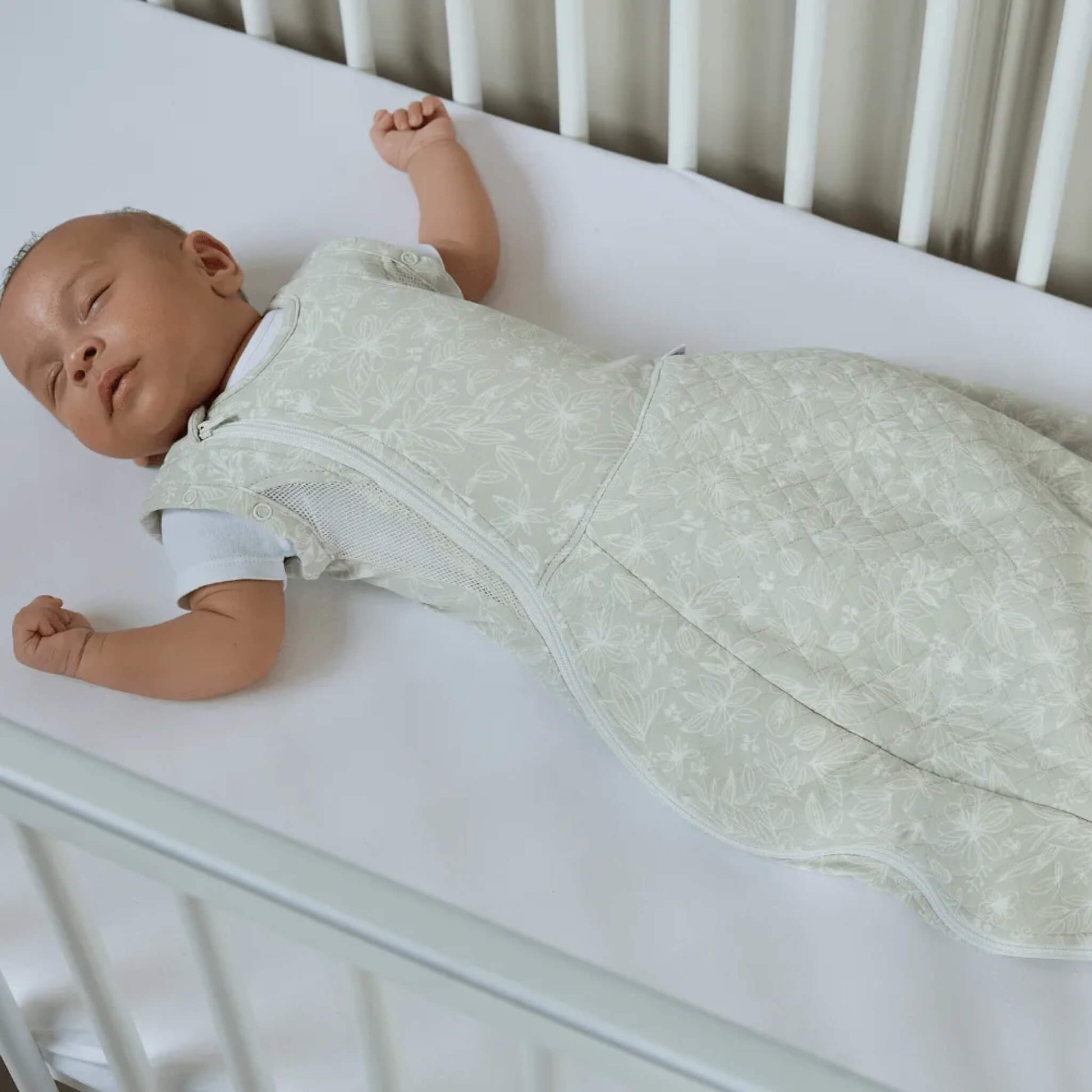 A baby lying in a cot wearing a swaddle sleep bag with arms positioned out, showing the natural sleep bag shape.