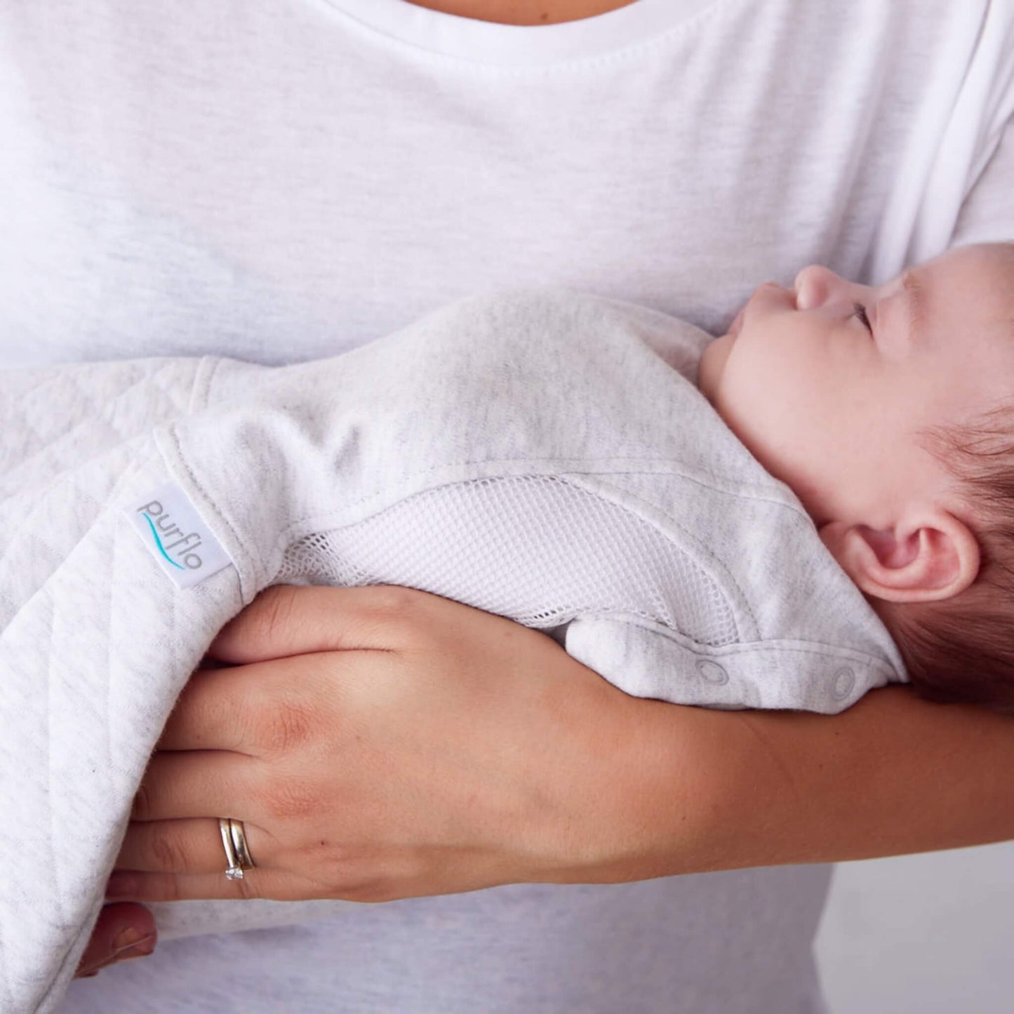 A baby resting against a mother’s chest while wearing a minimal grey swaddle sleep bag, showing breathable mesh panels along the sides.