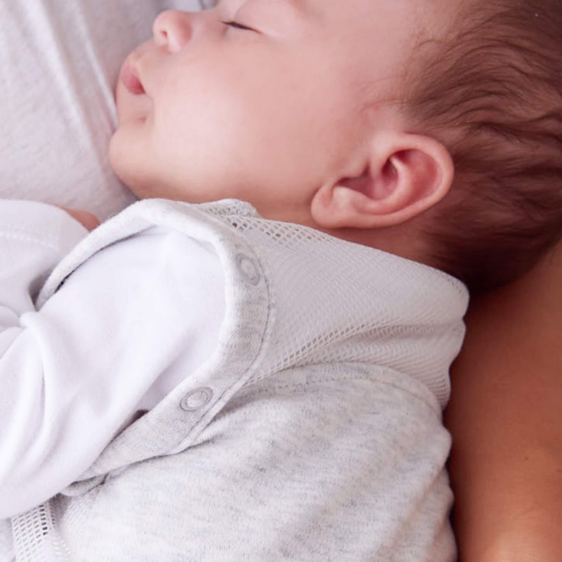 A close-up of a sleeping baby’s head and shoulders showing breathable mesh fabric at the neck and shoulder area of a light grey swaddle to sleep bag.