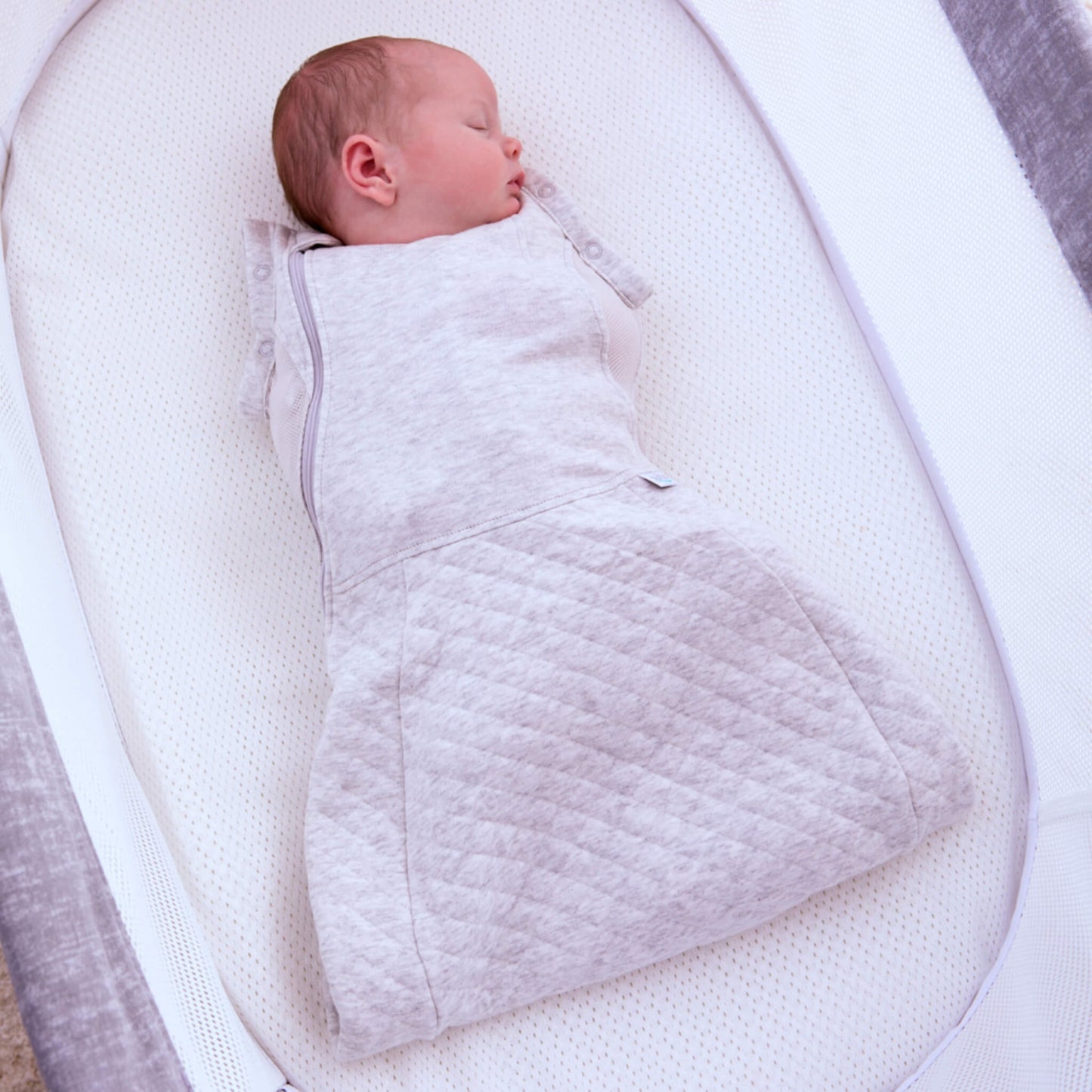 A baby sleeping on a breathable mattress while wearing a minimal grey swaddle sleep bag with arms secured inside for a snug swaddled feel.