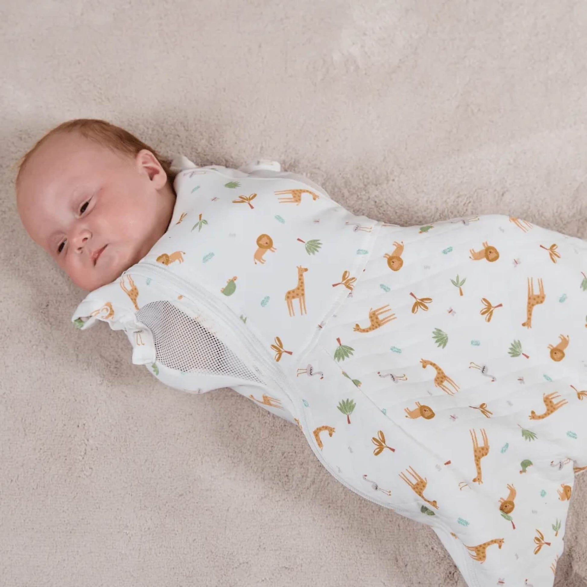 A baby lying on soft brown blanket wearing a swaddle to sleep bag, showing a fitted upper body and a roomy, padded lower section for leg movement.