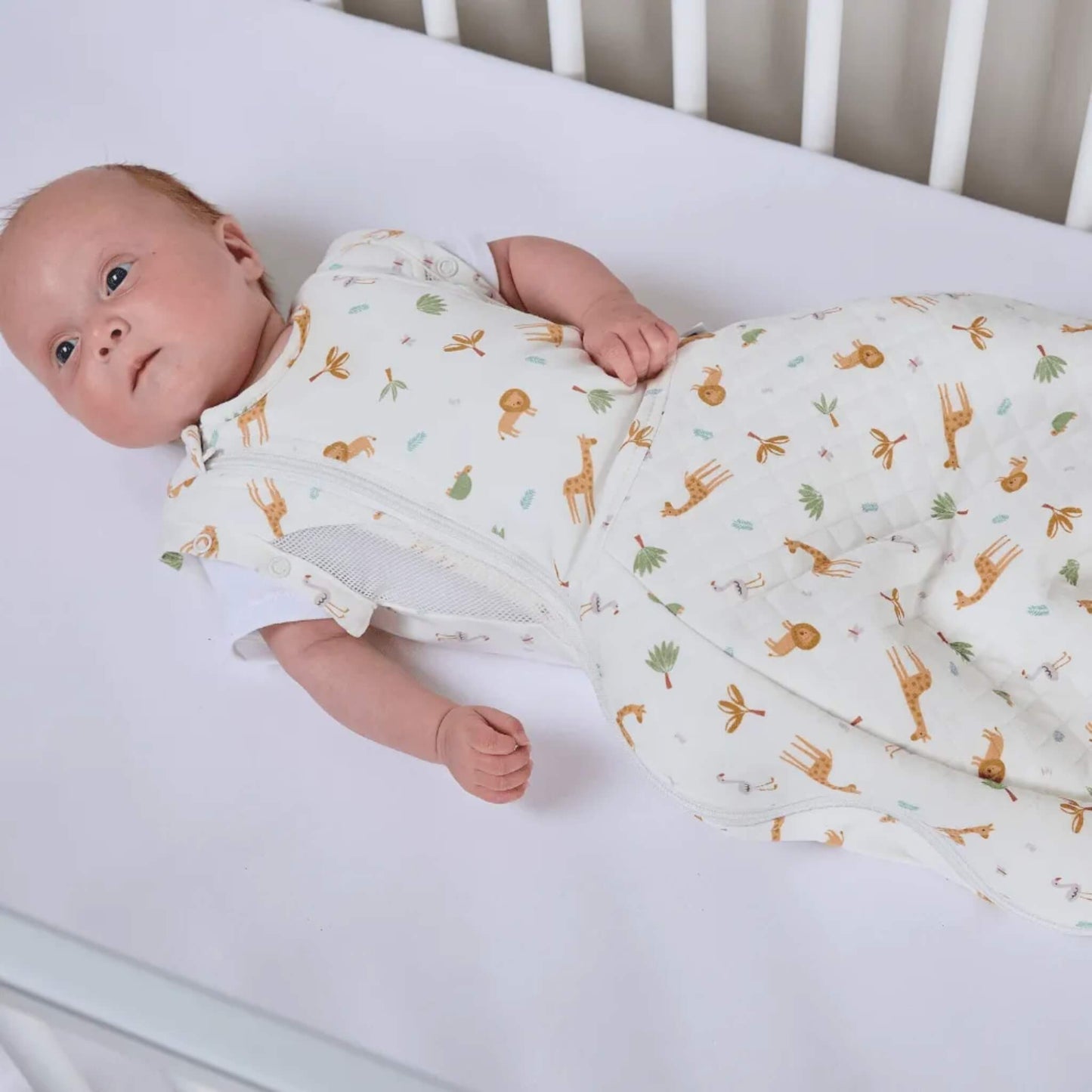 A baby lying on a soft surface wearing a swaddle to sleep bag with a safari animal print, showing arm-free sleeping with a zip-fastened front.