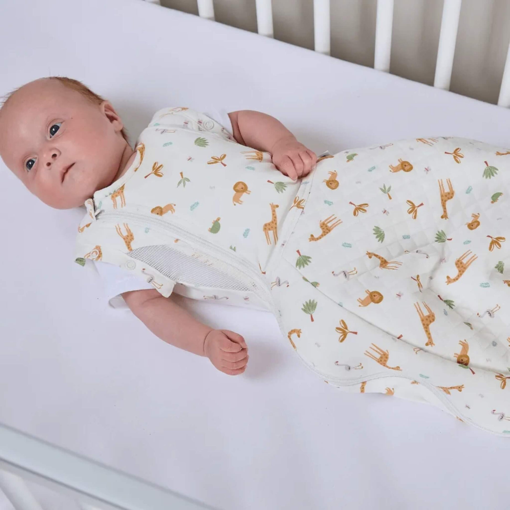 A baby lying on a soft surface wearing a swaddle to sleep bag with a safari animal print, showing arm-free sleeping with a zip-fastened front.