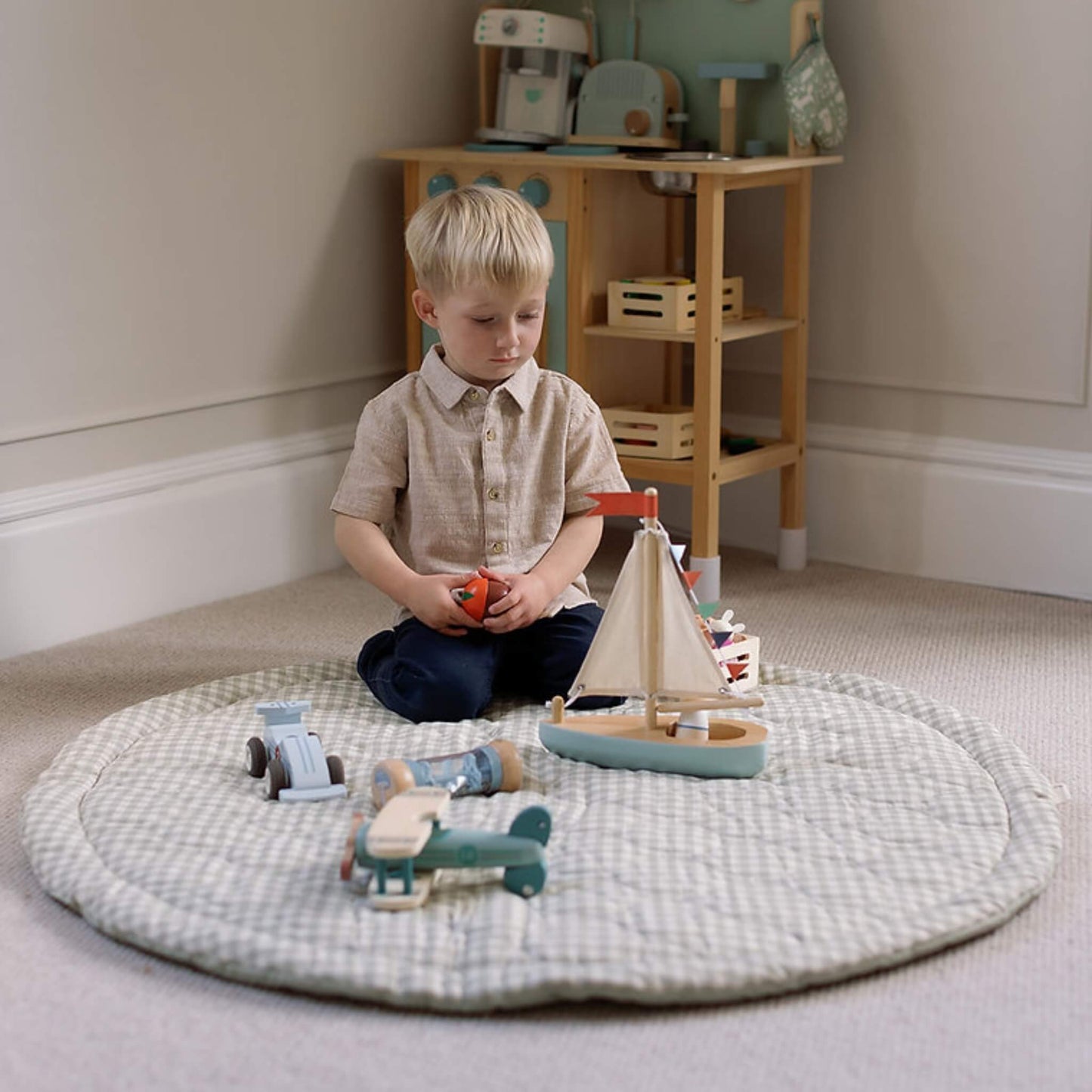A young child seated on a quilted floor mat in a neutral room, surrounded by wooden toys including a sailboat, car and stacking pieces.