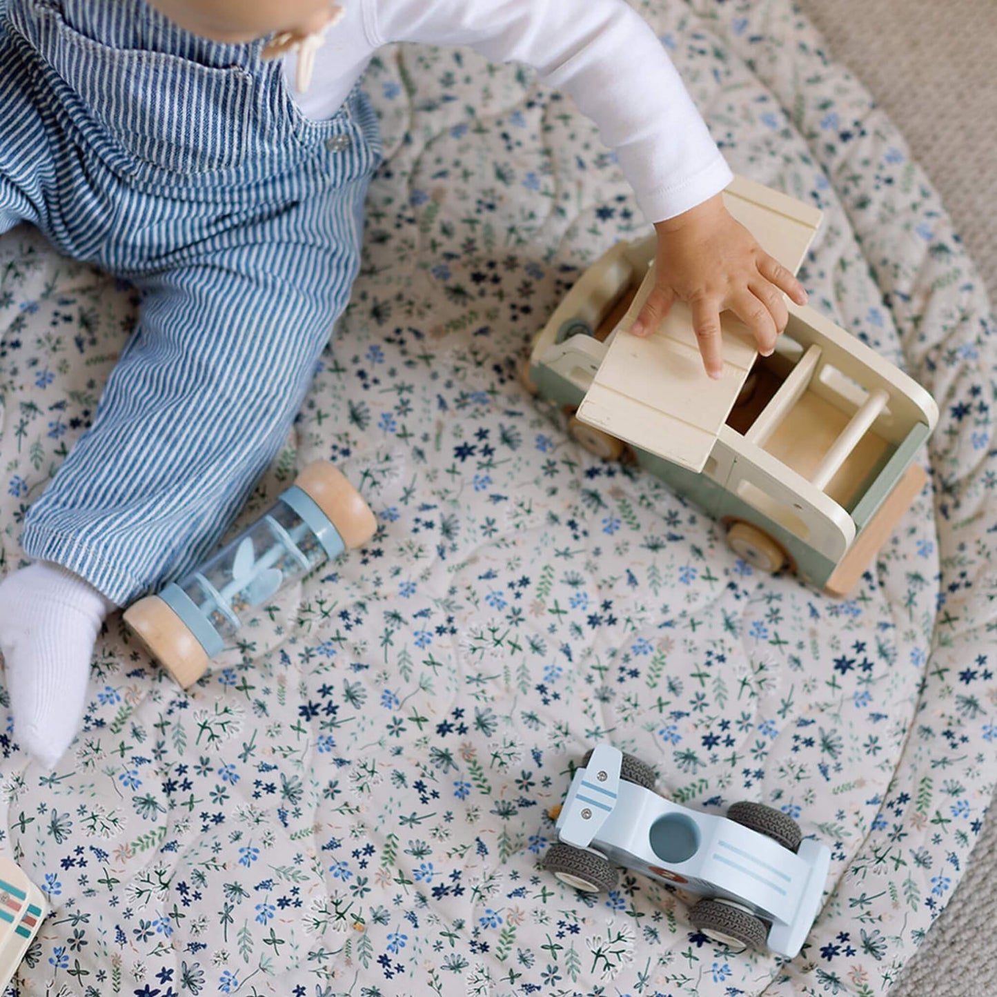 A close-up of a baby’s hand opening a wooden toy truck on a quilted play mat, showing the soft padded texture and detailed stitching underneath.