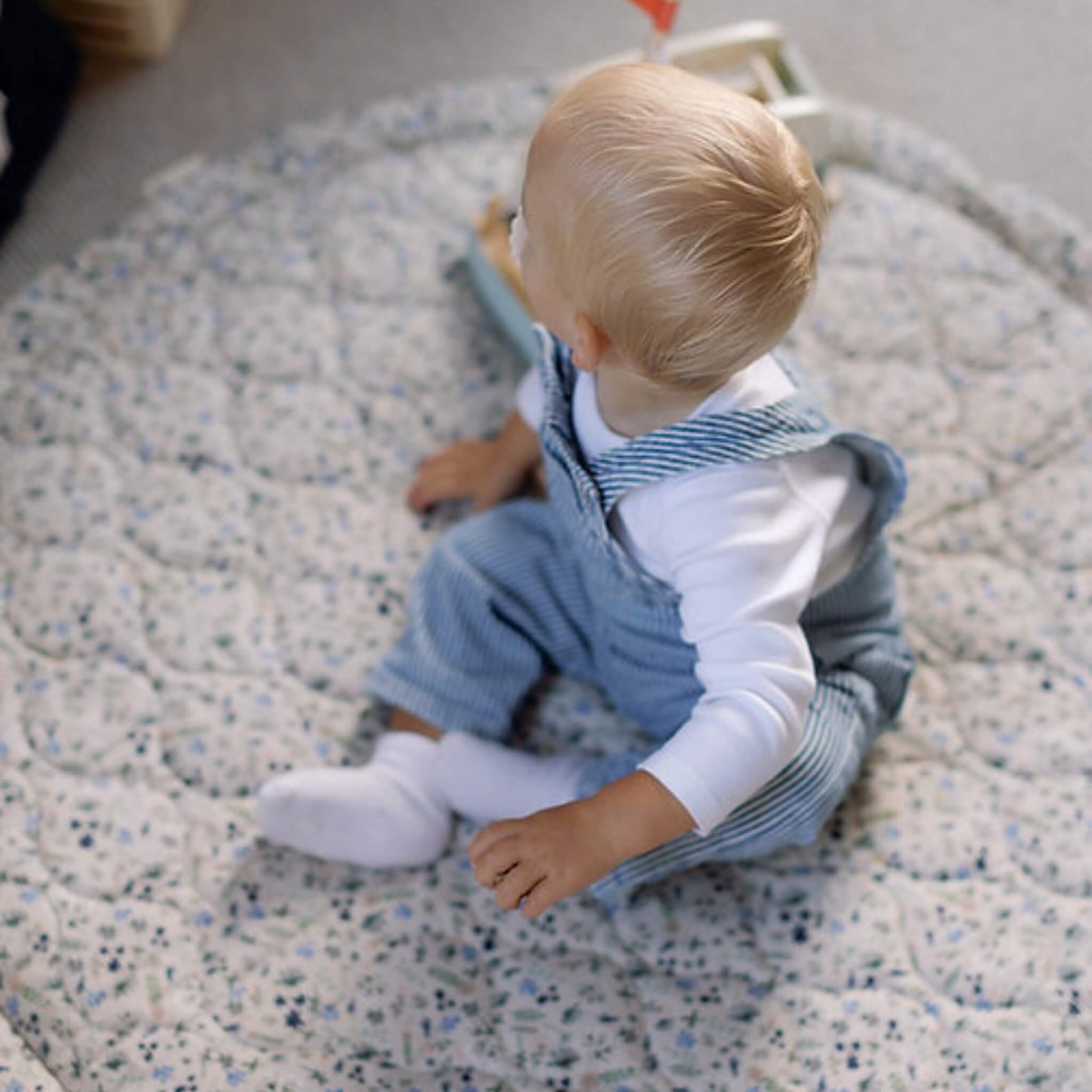 A baby sitting on a quilted floor play mat looking behind, showing the padded surface and gentle cushioning.