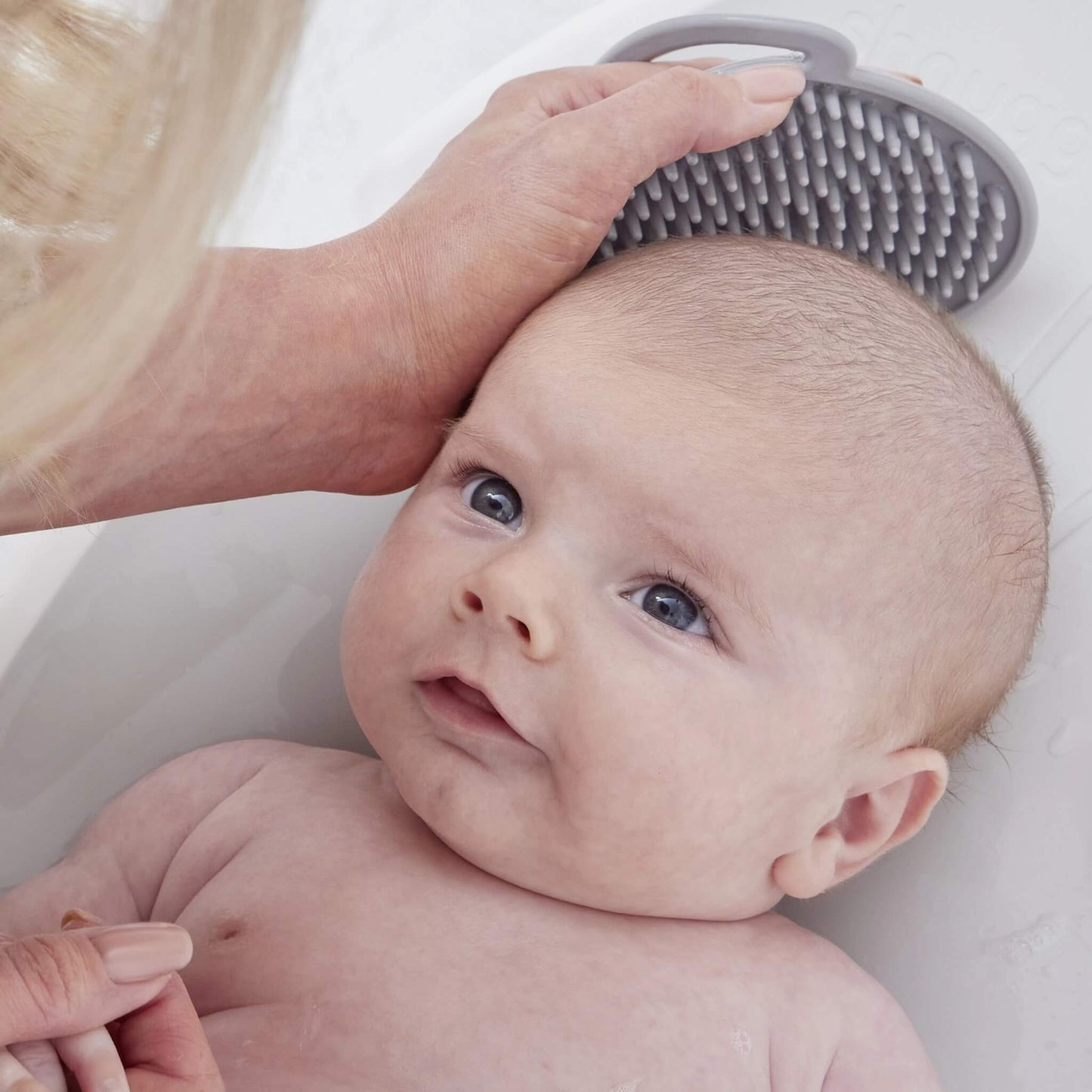 A baby lying in a bath while an adult uses a soft silicone brush to gently clean a baby’s scalp.