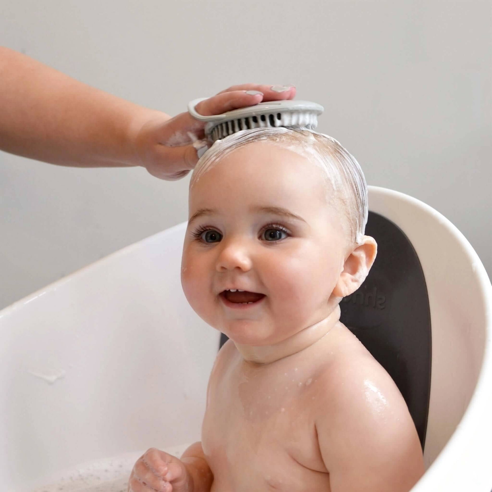 A smiling baby sitting in a bath while hair is washed using a soft silicone scalp brush.