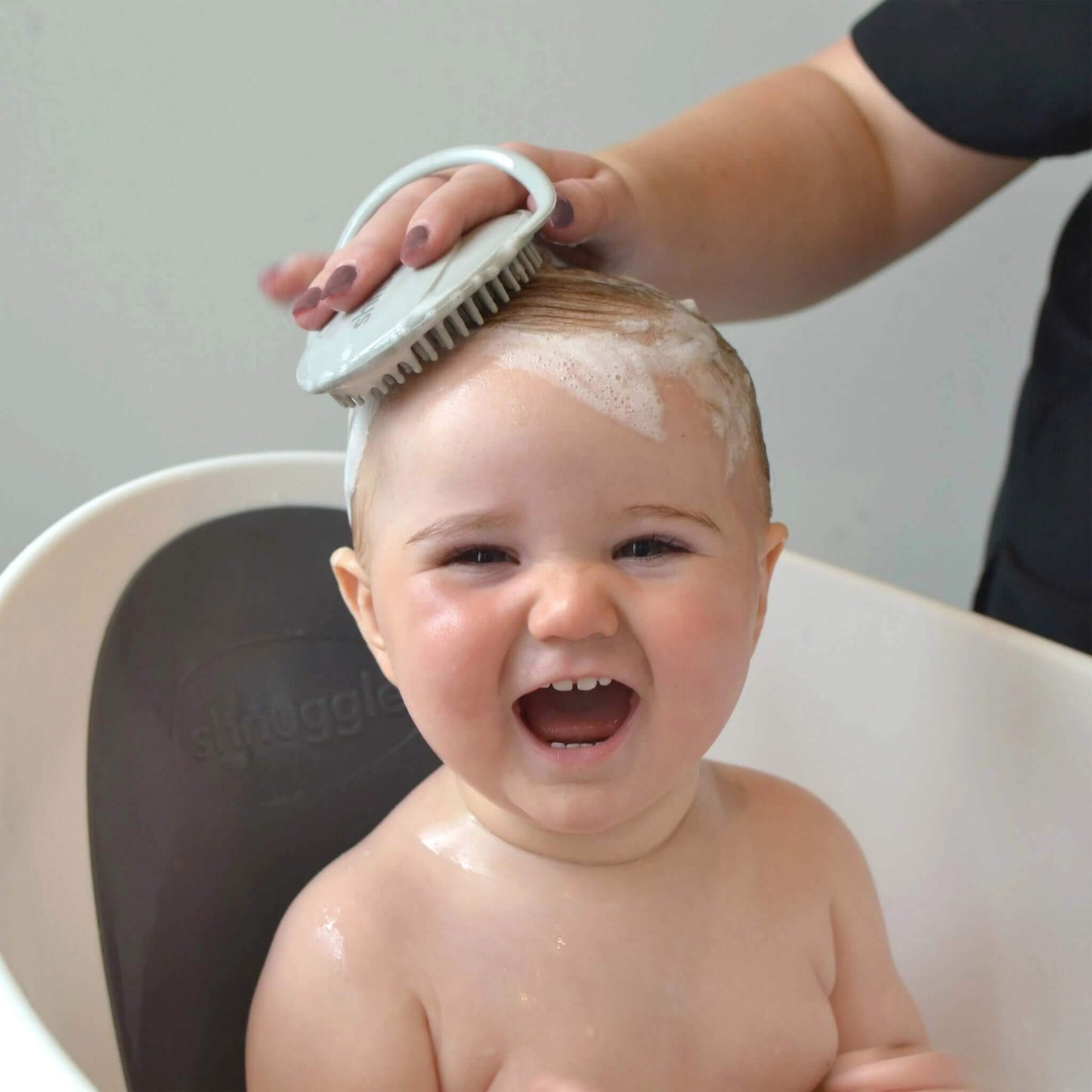 A mother gently washing a baby’s hair in a bath using a soft silicone scalp brush, with light lather visible and the baby smiling comfortably.