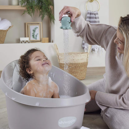 A toddler sitting in a grey baby bath, smiling as water pours down from a mint green octopus bath toy held above.