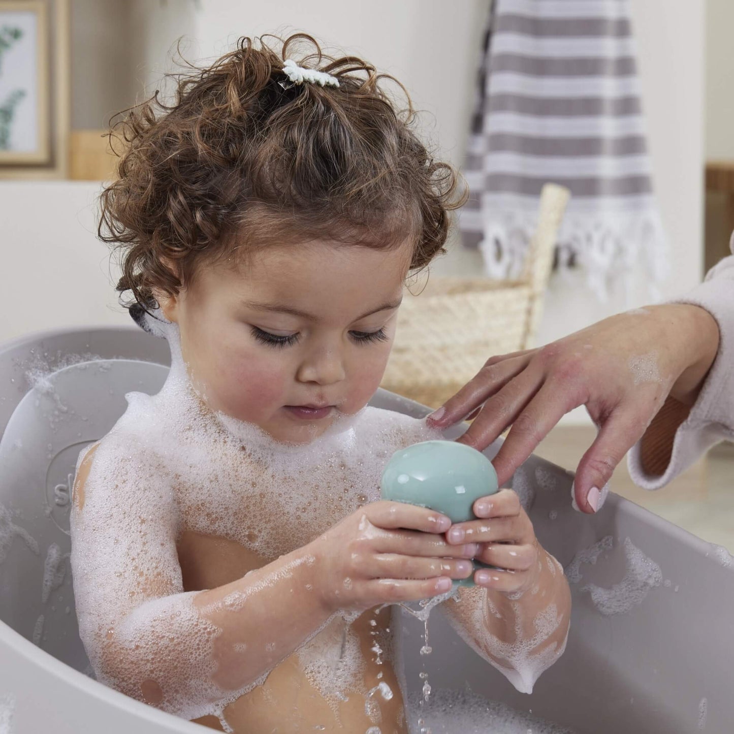 A toddler in a bubble bath holding a mint green octopus bath toy while an adult supports them gently.