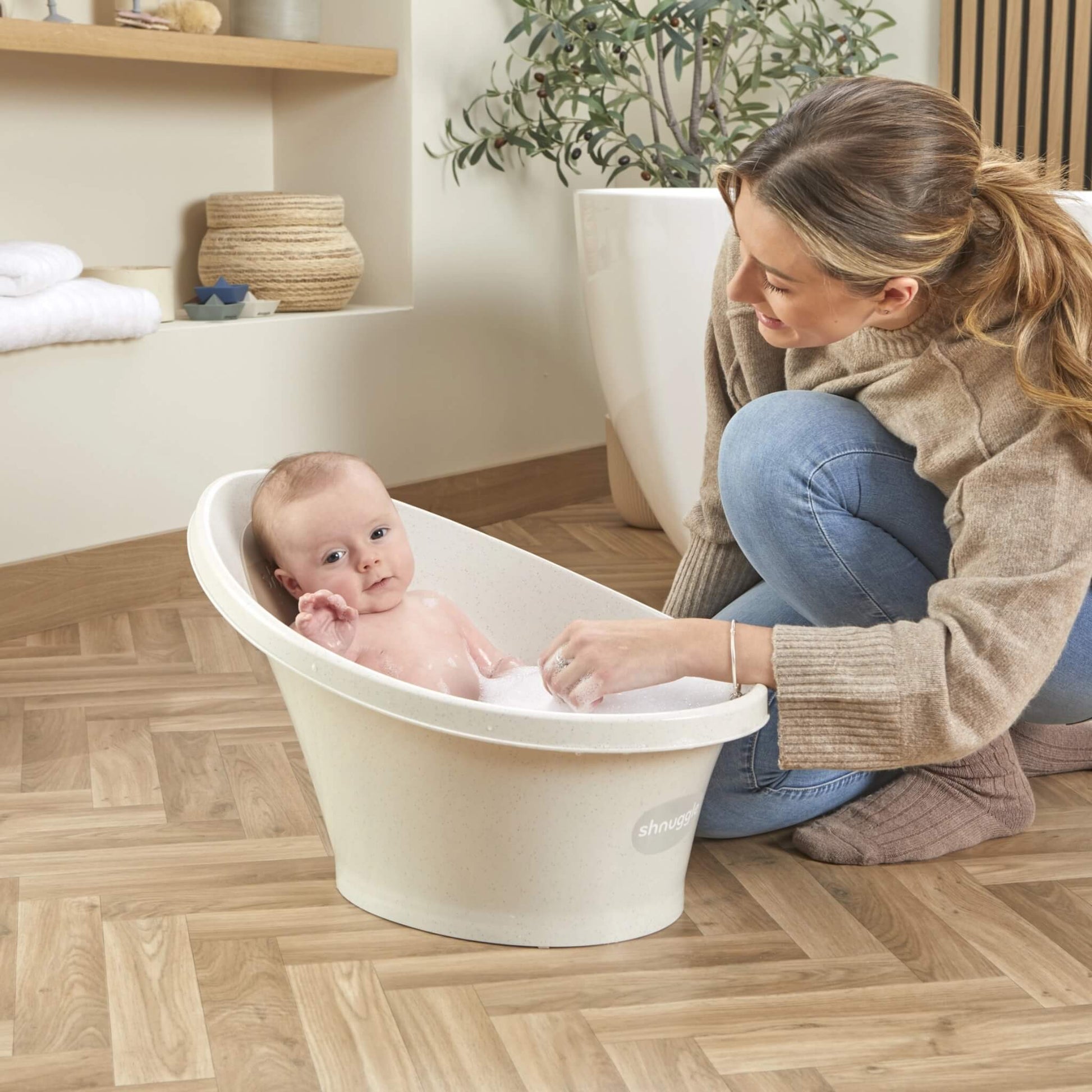 Baby seated in a bath with bubbles, looking up while their mother kneels beside them.