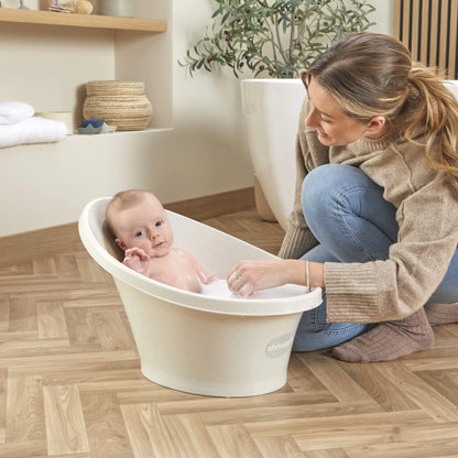 Baby seated in a bath with bubbles, looking up while their mother kneels beside them.