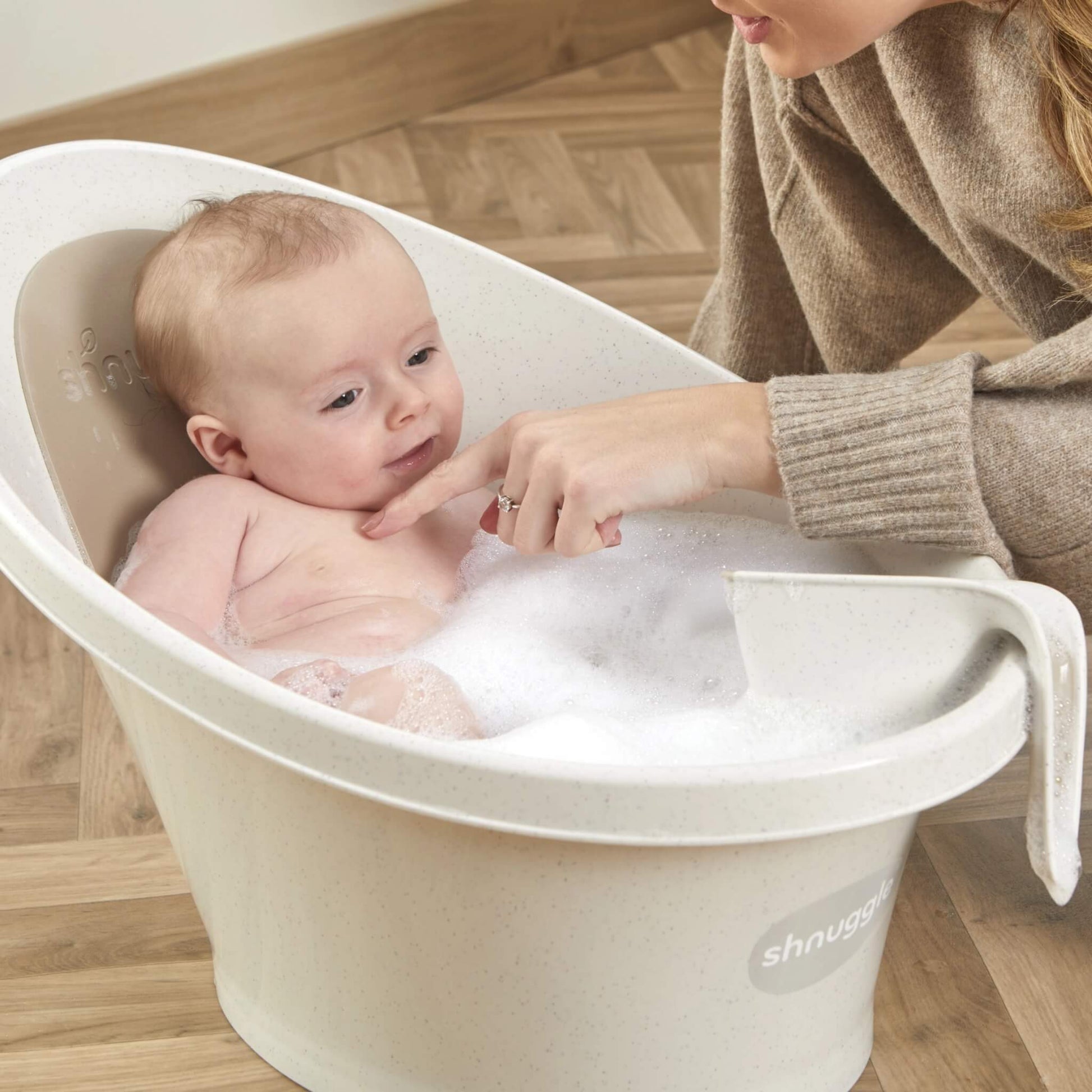 Smiling baby reclining in a bubble bath while their mother touches the baby’s chin with a finger.