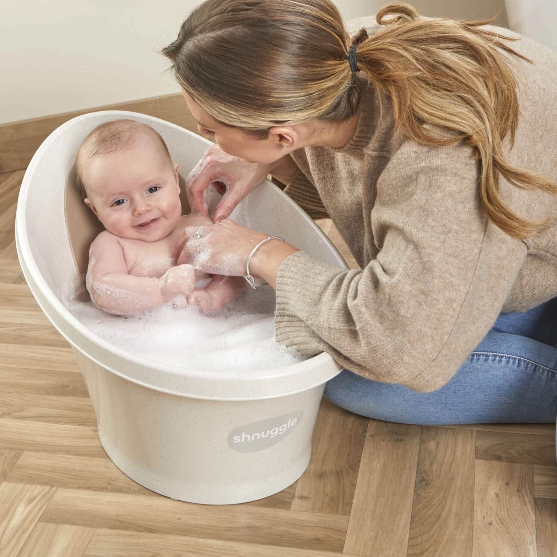 Baby in a bubble-filled bath smiling as their mother leans forward and gently washes them