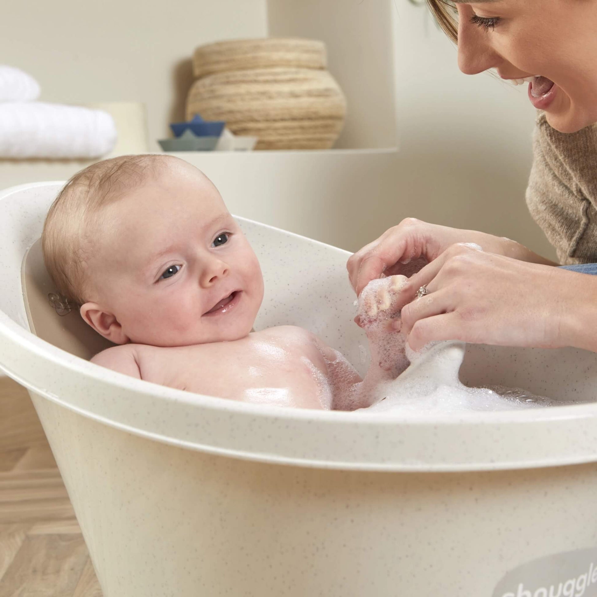 Baby laughing in a bath of bubbles while their mother holds their hand and plays during washing.
