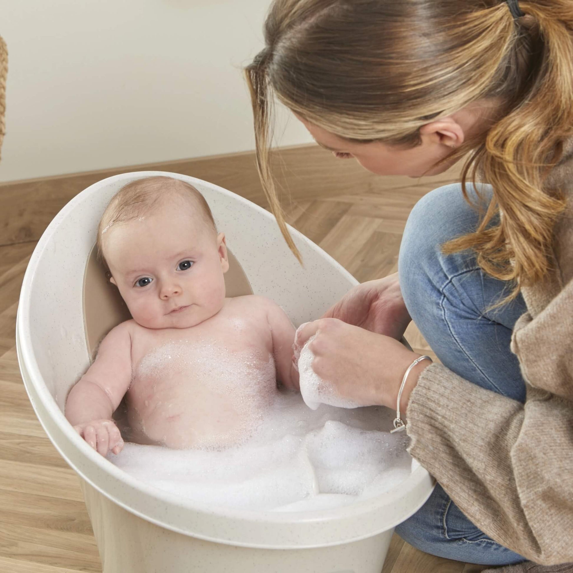 Mother washing her baby who is seated upright in a small bath filled with bubbles.