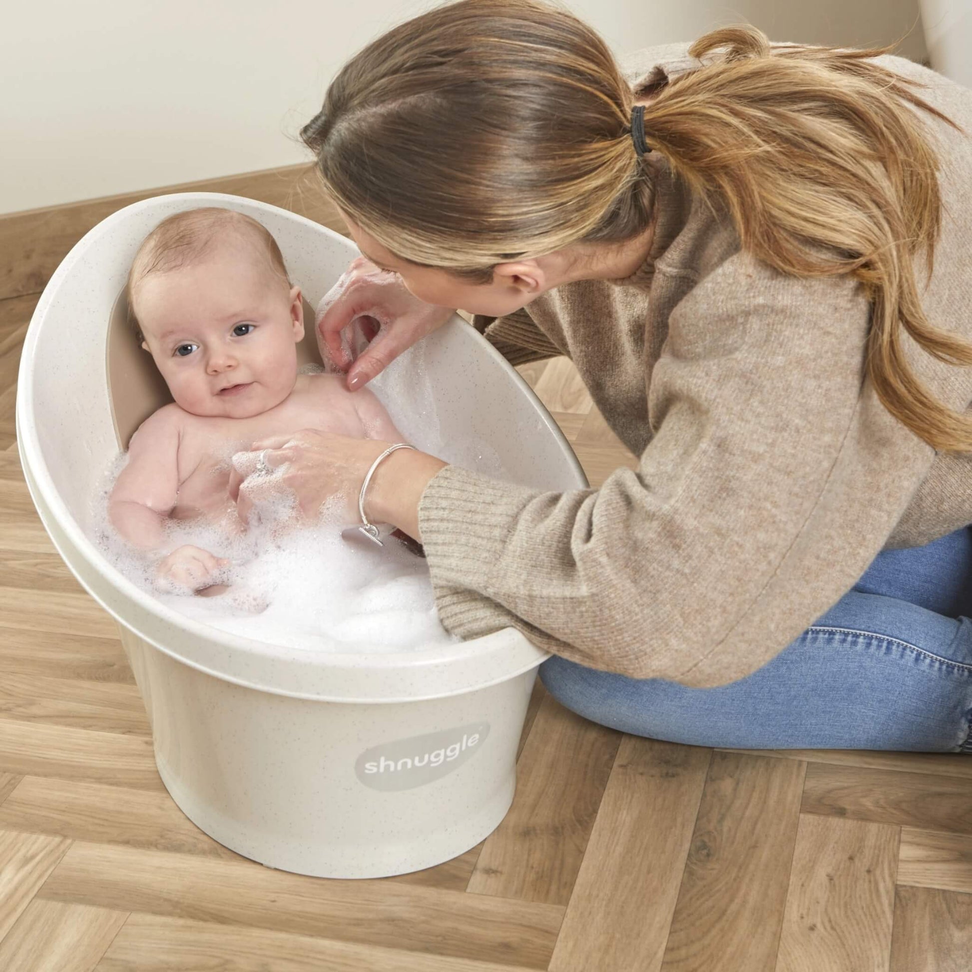 Smiling baby sitting in a bubble-filled bath as their mother gently rinses them.