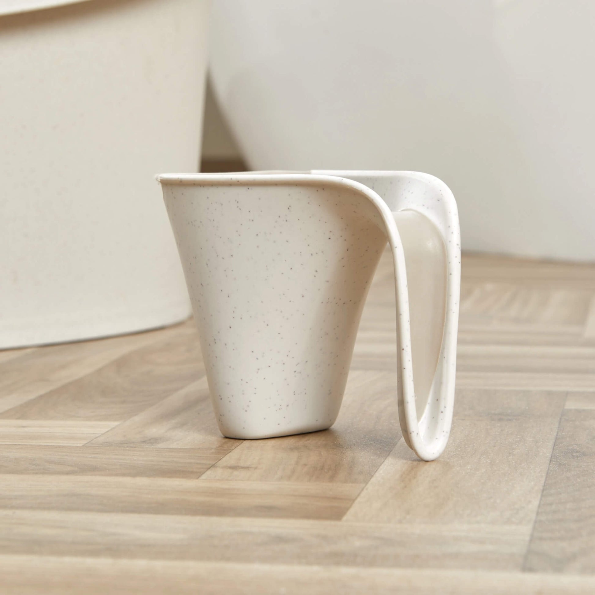 Side view of a cream jug with speckled finish and wide handle, standing on a wooden floor.