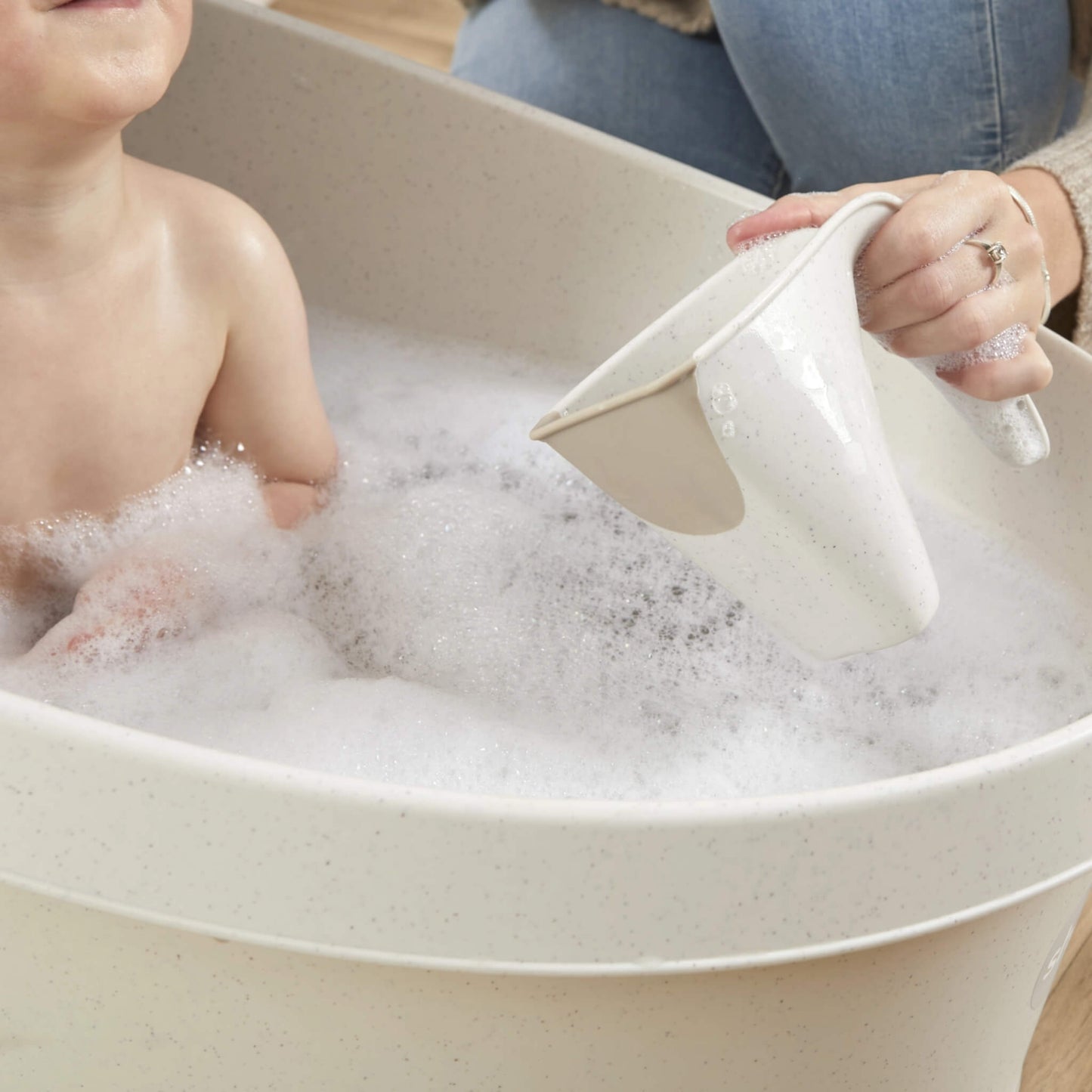 Hand holding a cream jug above a baby in a bubble bath, pouring water gently over their skin.