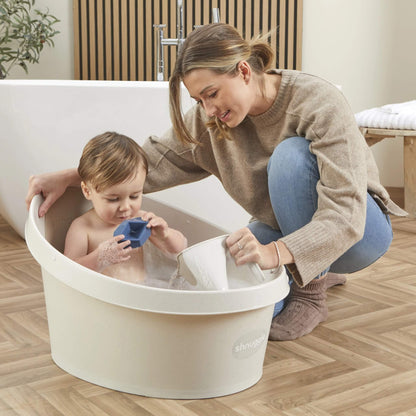 Mother bathing a toddler while using a cream jug to rinse, with the child sitting in a bubble bath and playing with a toy boat.