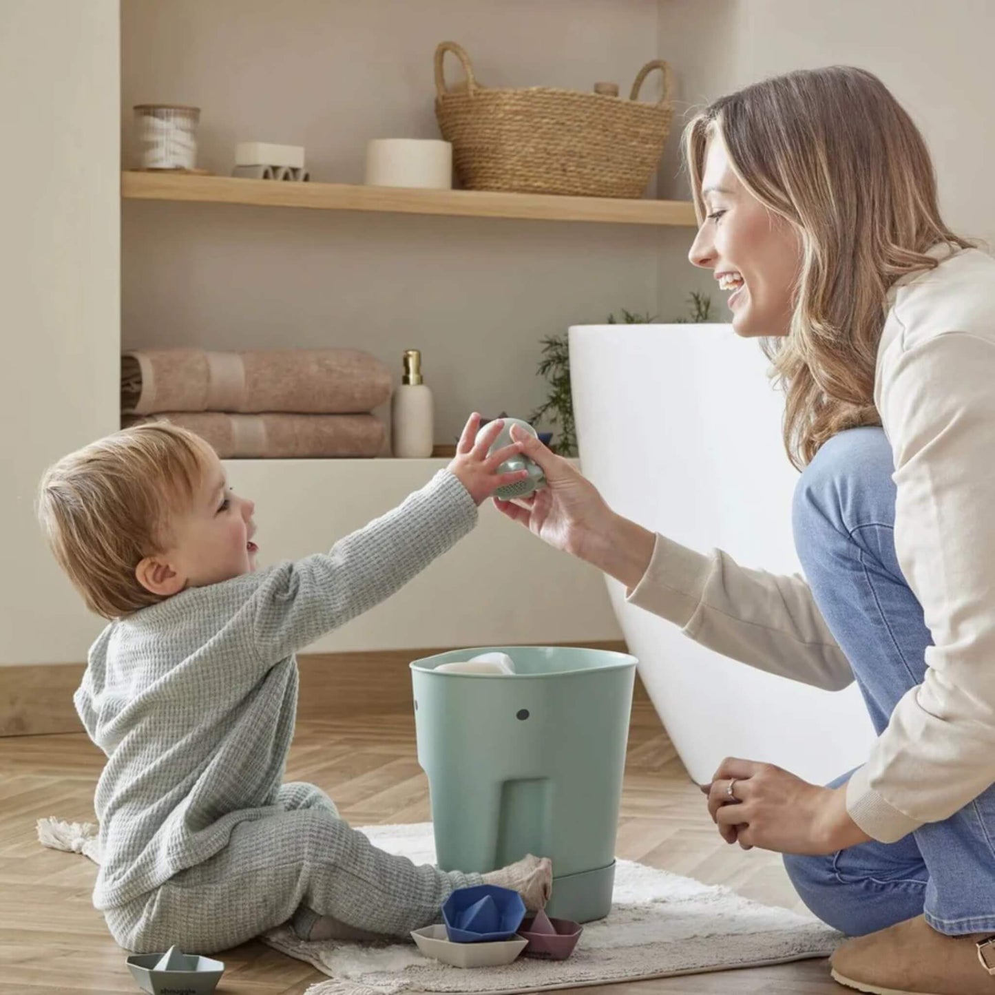 A toddler sitting on a mat hands a bath toy to a smiling mother, with a green elephant-shaped toy caddy beside them indoors.