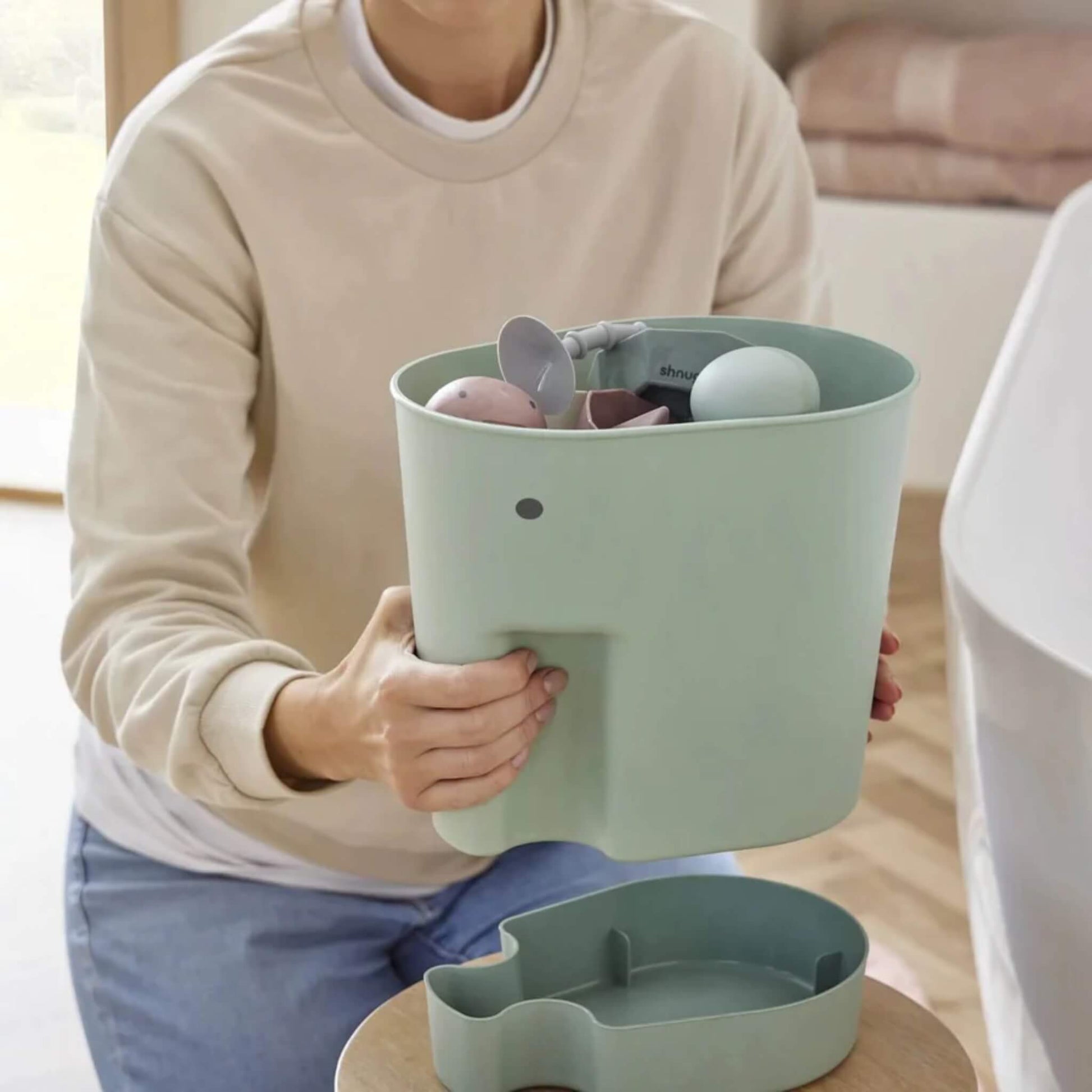 A mother lifts a green elephant-shaped toy caddy filled with bath toys above a matching drip tray placed on a stool.