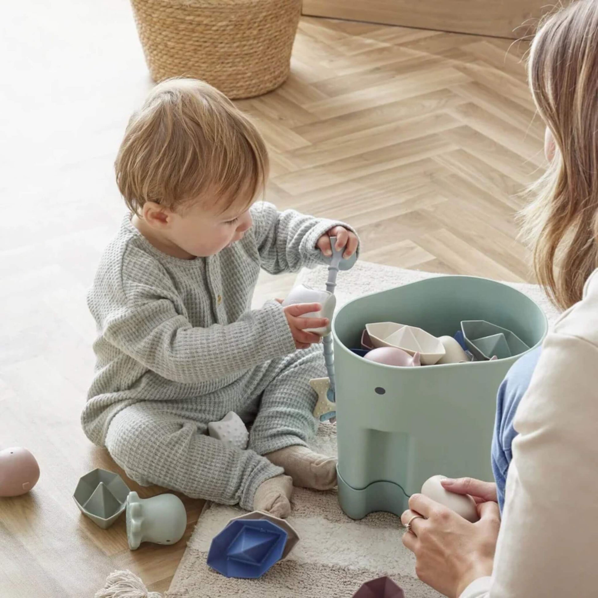A toddler sits on a mat placing bath toys into a green elephant-shaped caddy while a mother kneels nearby.