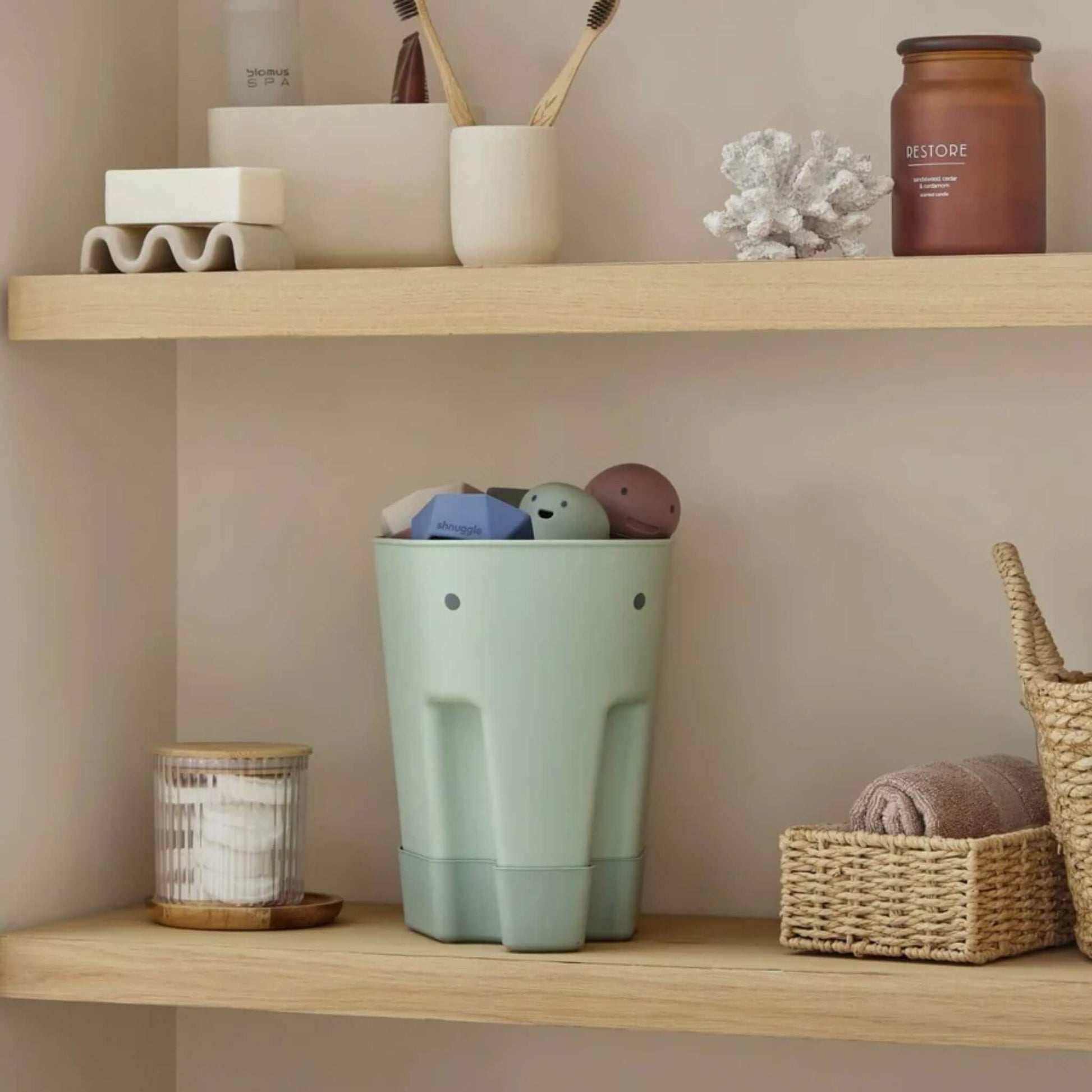 A green elephant-shaped bath toy caddy stands on a wooden shelf holding assorted bath toys in a calm bathroom setting.