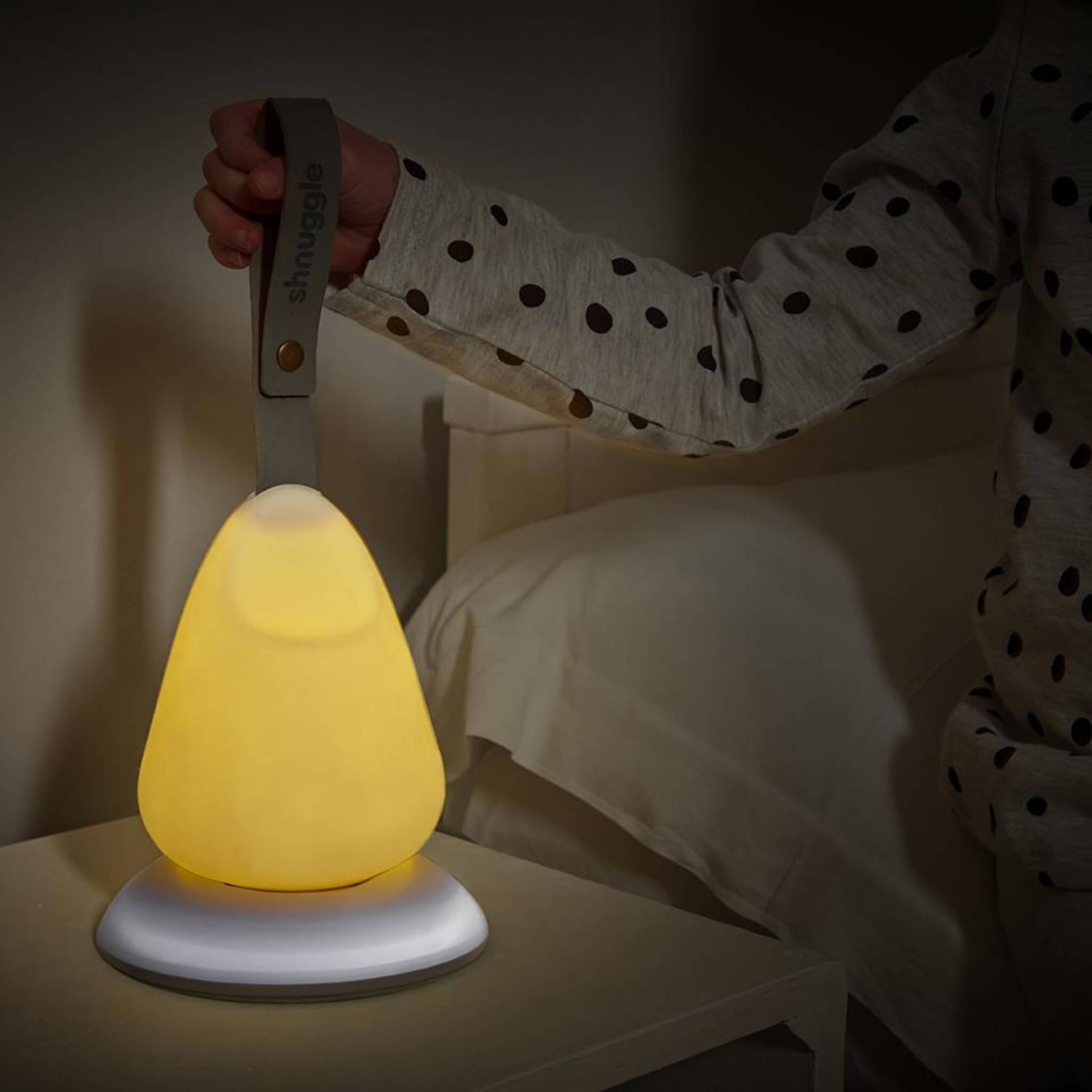 Close-up of a child’s hand holding the strap of a portable nightlight placed on a bedside table, glowing with a warm yellow light.