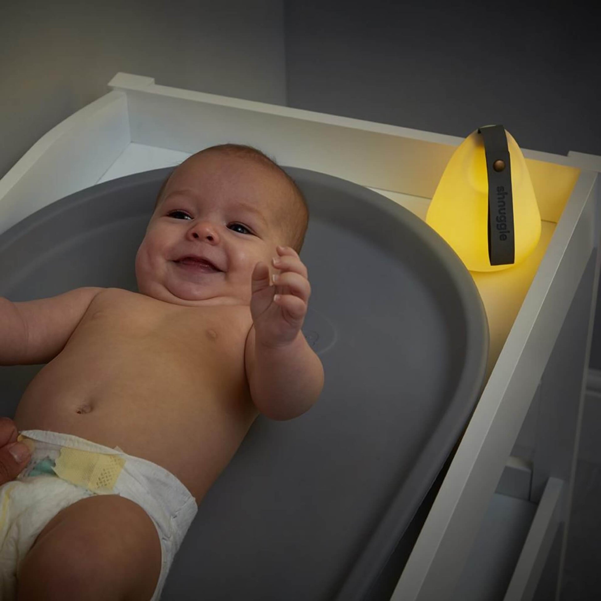 Smiling baby lying on a grey changing mat with a glowing nightlight nearby, providing soft illumination for nighttime nappy changes.