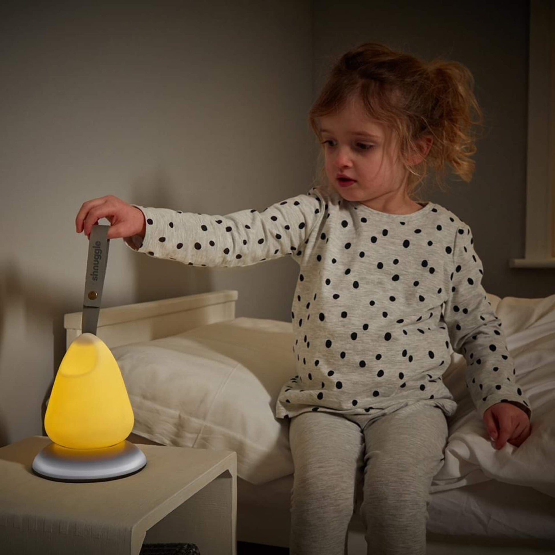 Young child sitting on a bed, reaching to switch on a glowing nightlight placed on a bedside table.