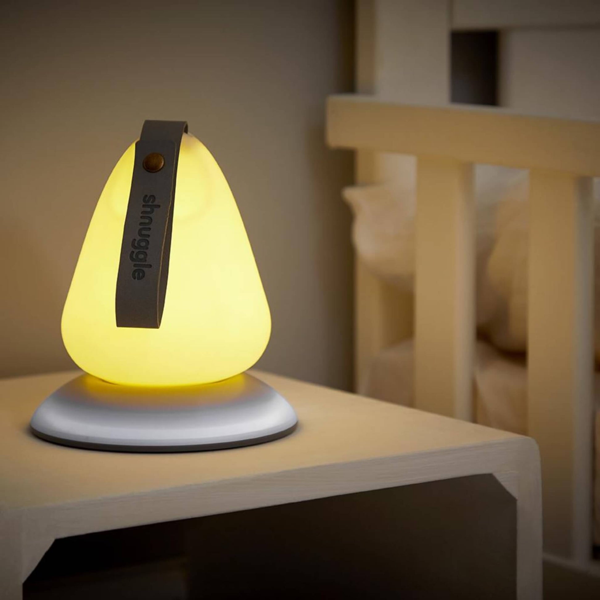 Nursery nightlight glowing warmly on a small table beside a cot in a dimly lit room.