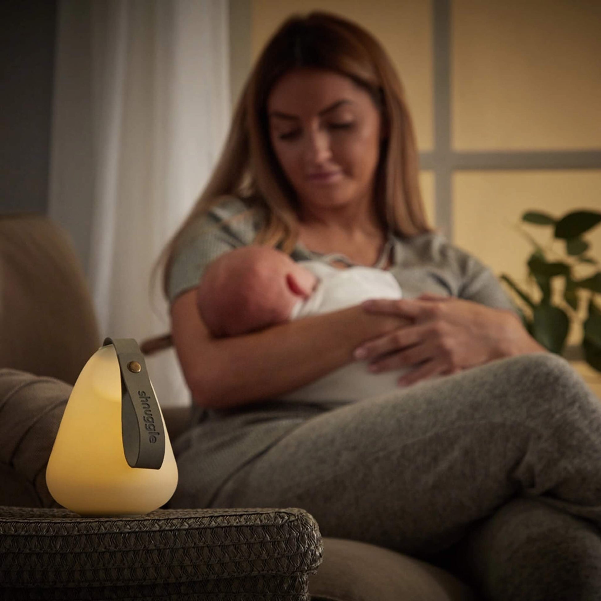 Mother sitting on a chair holding her baby, with a glowing nightlight resting on the arm of the chair in the foreground.