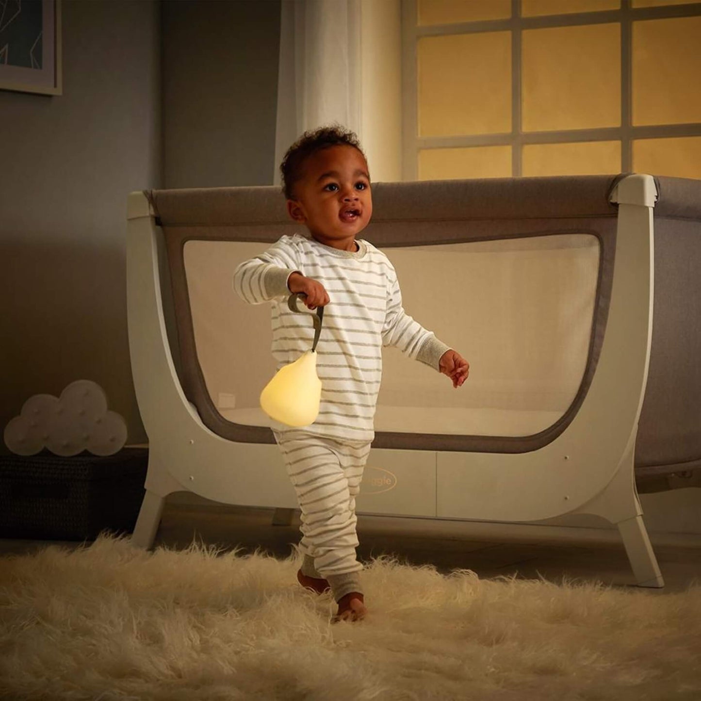 Smiling toddler in striped pyjamas holding a glowing portable nightlight by its strap while walking across a nursery with a cot in the background.