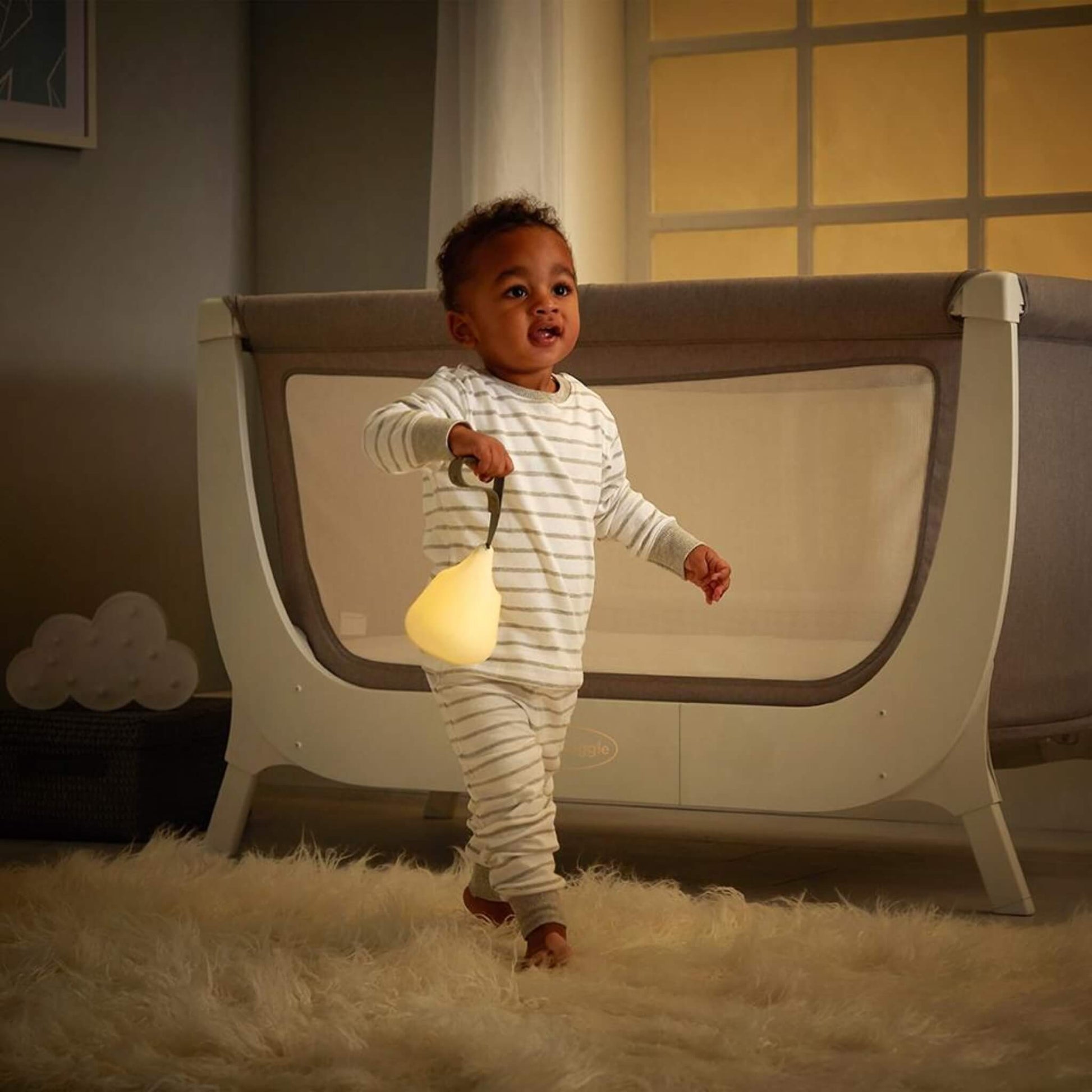 Smiling toddler in striped pyjamas holding a glowing portable nightlight by its strap while walking across a nursery with a cot in the background.
