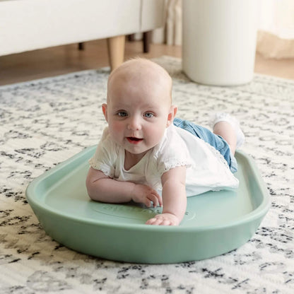 A baby lying on their tummy on a eucalyptus green foam changing mat placed on a patterned rug in a living room setting.