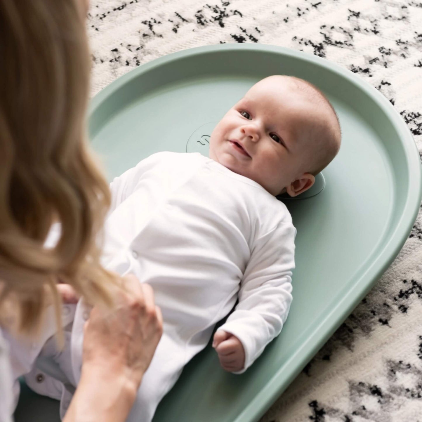 A baby lying on their back on a contoured eucalyptus green changing mat while the mother fastens a sleepsuit, showing the raised supportive sides.