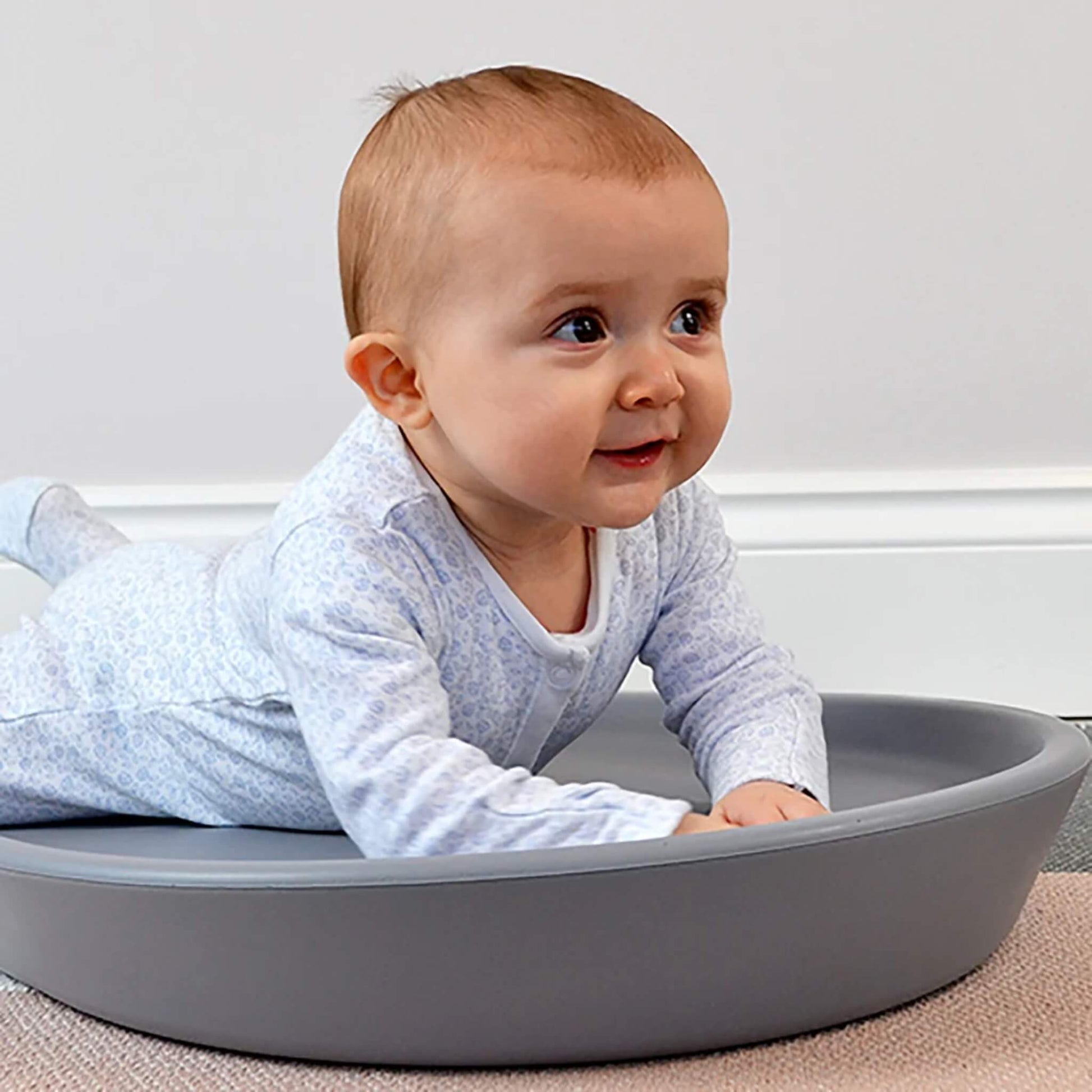 Changing Mat in grey with a baby lying on their tummy, showing the raised cushioned sides and contoured foam design on a carpeted floor.