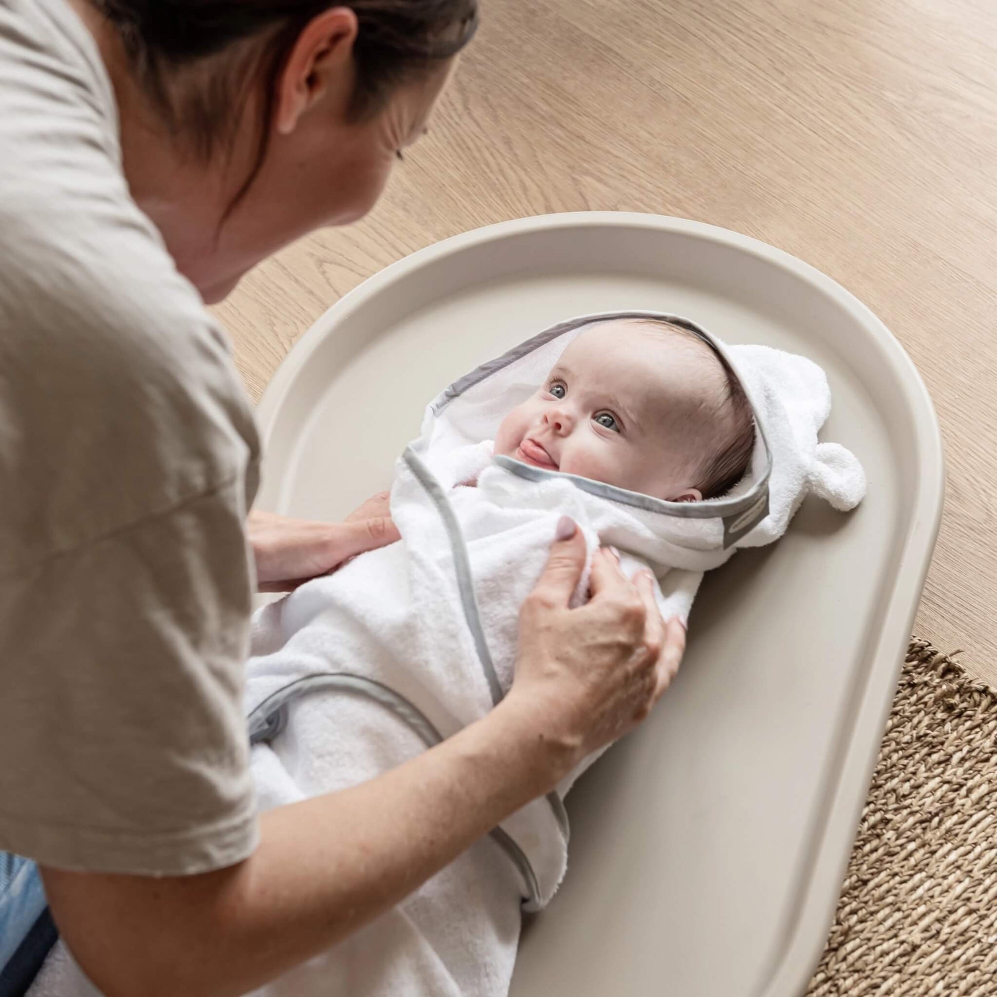 Mother gently wrapping her baby in a hooded towel while the baby lies on a taupe changing mat on a wooden floor.
